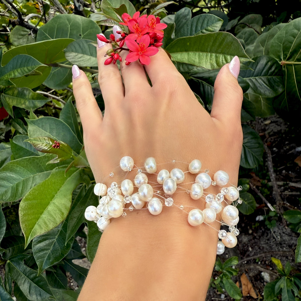 Elegant pearl and crystal bracelet worn on a woman's wrist while holding vibrant red flowers, with lush green foliage in the background.