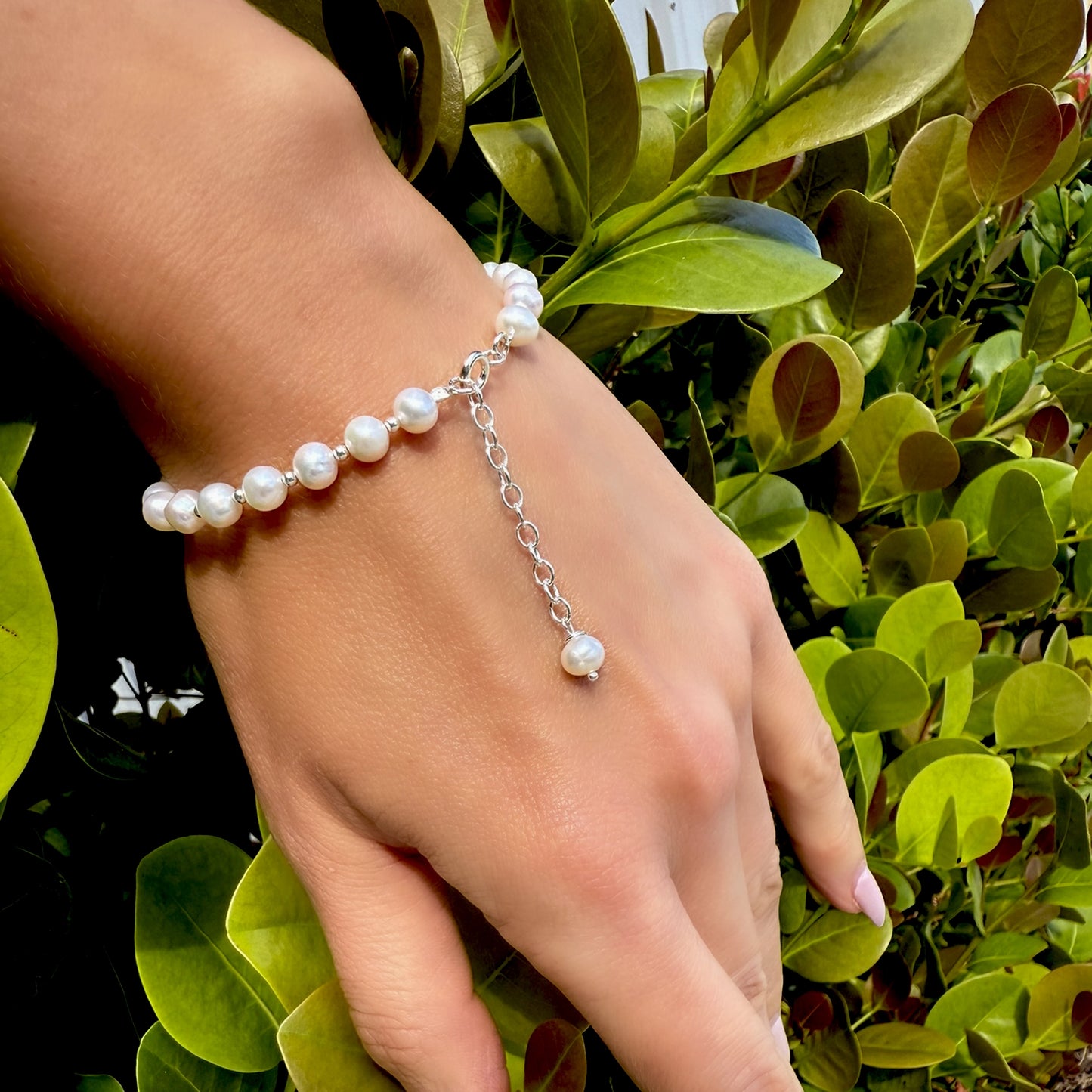 White freshwater pearl bracelet with silver adjustable chain extension, worn on wrist in front of vibrant green foliage.