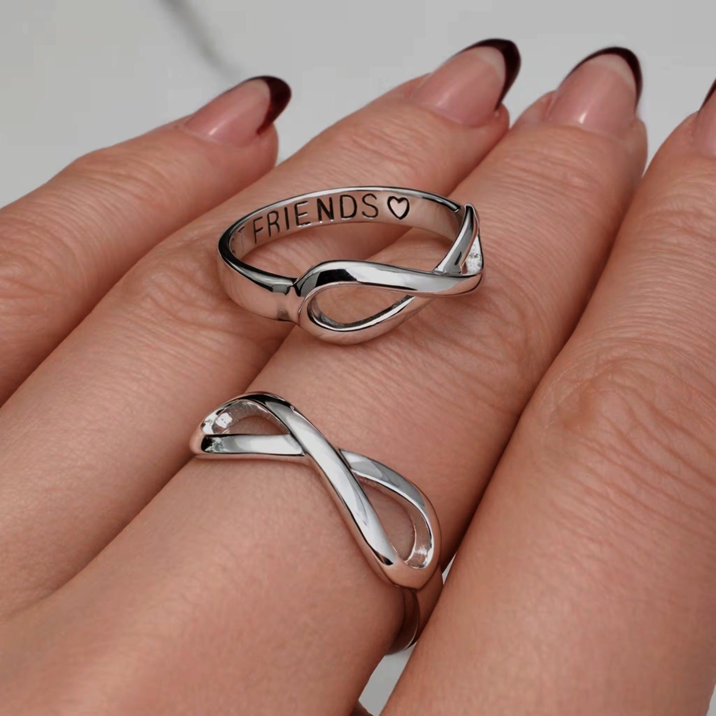 Silver infinity friendship rings on a woman’s hand, one engraved “FRIENDS” with a heart, polished and shiny with soft romantic lighting, perfect Valentine’s Day gift jewelry.