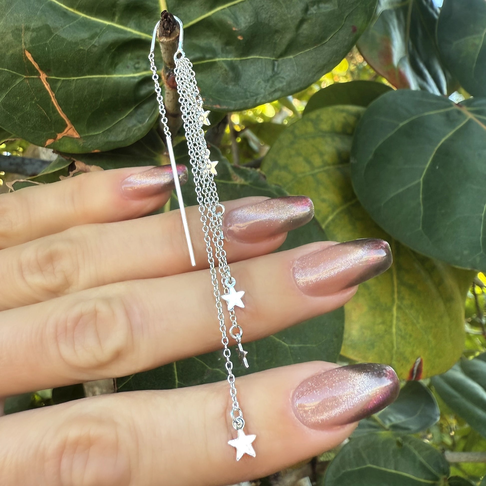 Close-up of a hand with glossy metallic pink gradient manicure holding a pair of sterling silver threader earrings. The earrings feature delicate dangling chains adorned with small star charms, showcased against a background of large green leaves.