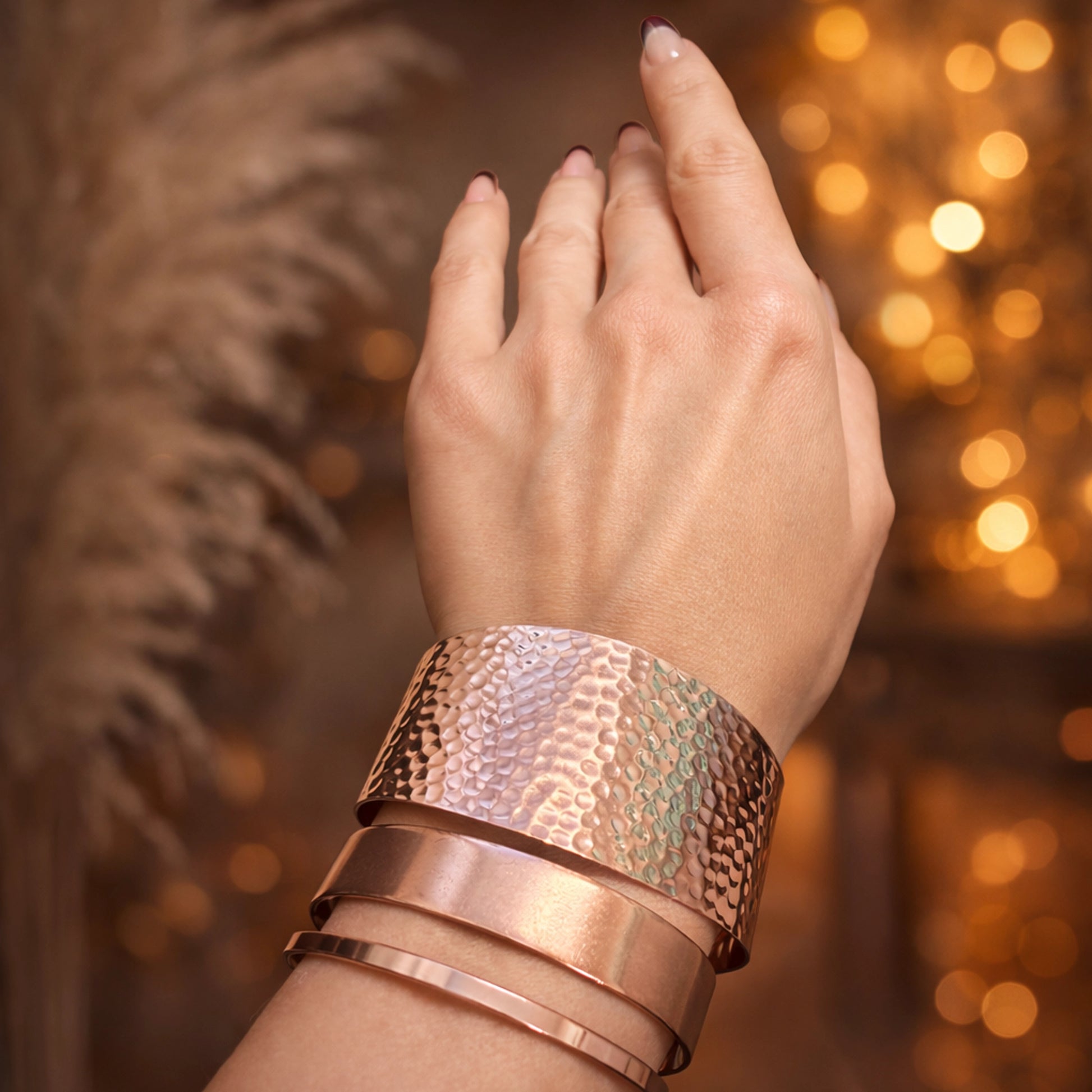 Wide hammered copper cuff bracelet stacked with minimalist copper bangles on a woman’s wrist, warm golden bokeh background, artisanal handcrafted jewelry, elegant boho style.
