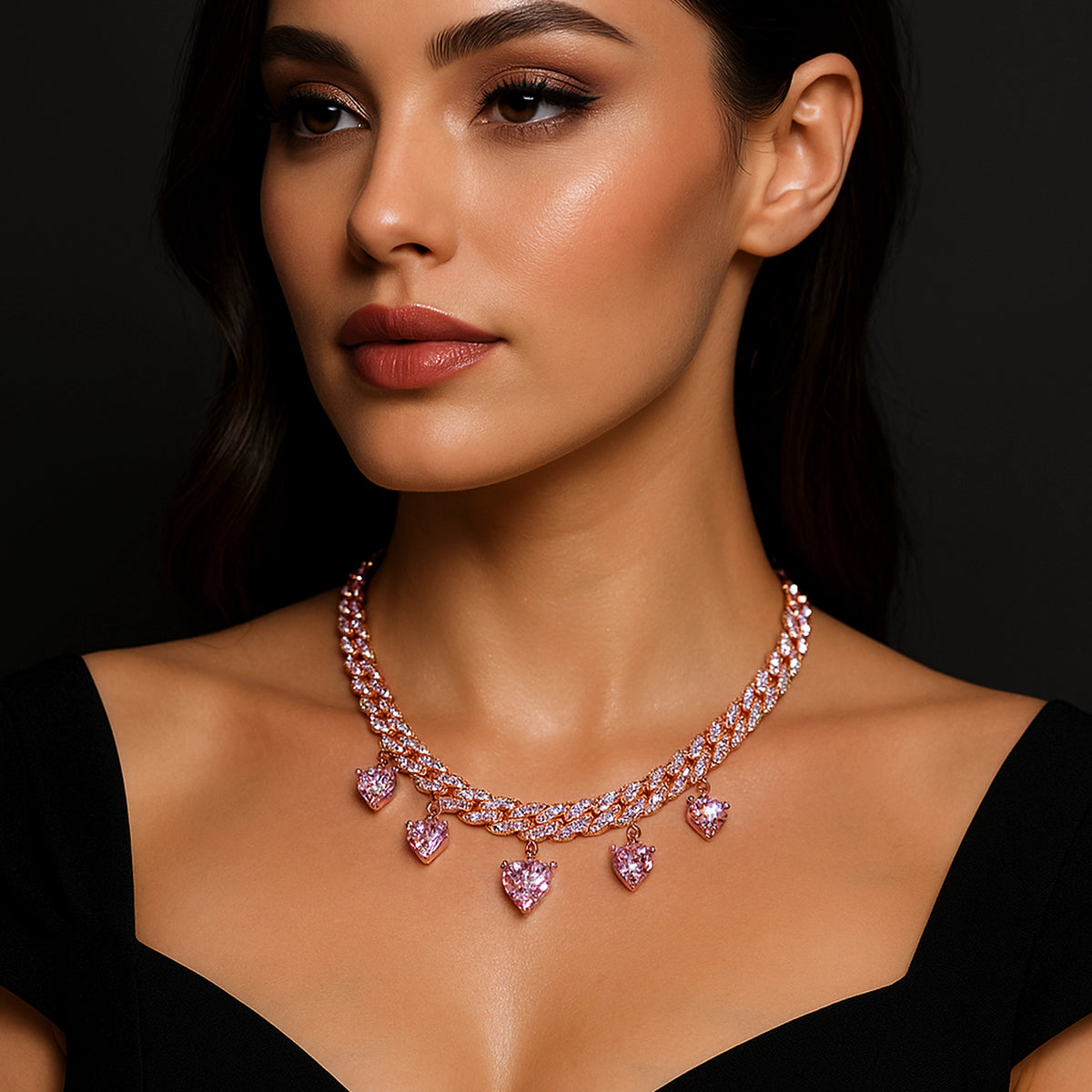 Close-up of a woman in a black off-shoulder dress wearing a statement rose gold curb chain necklace featuring sparkling pink heart-shaped CZ charms and shimmering pavé links.