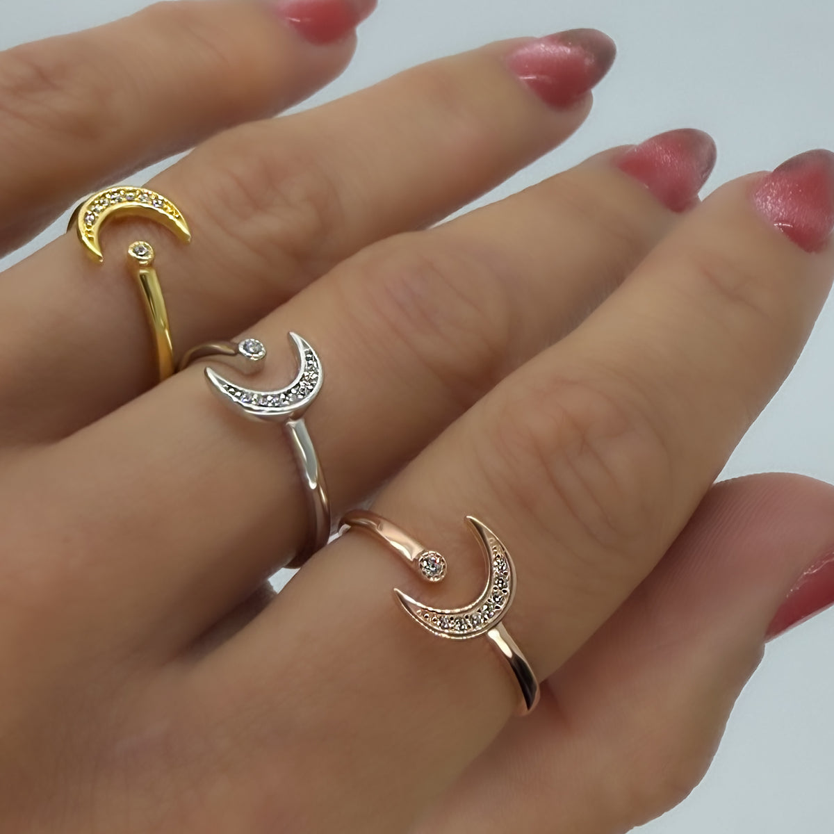 Close-up of three fingers wearing crescent moon and star rings in yellow gold, sterling silver, and rose gold finishes. Each open ring features a sparkling CZ-studded moon and a tiny star accent, perfect for stacking or gifting as dainty celestial jewelry.