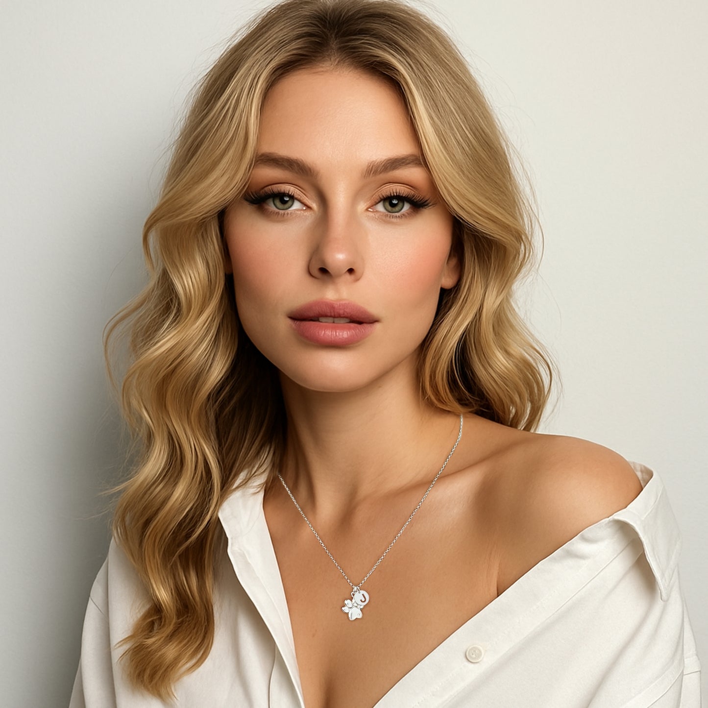 Blonde woman with soft waves and natural makeup wearing a white off-shoulder shirt, showcasing a sterling silver necklace with a CZ paw print charm and initial pendant.
