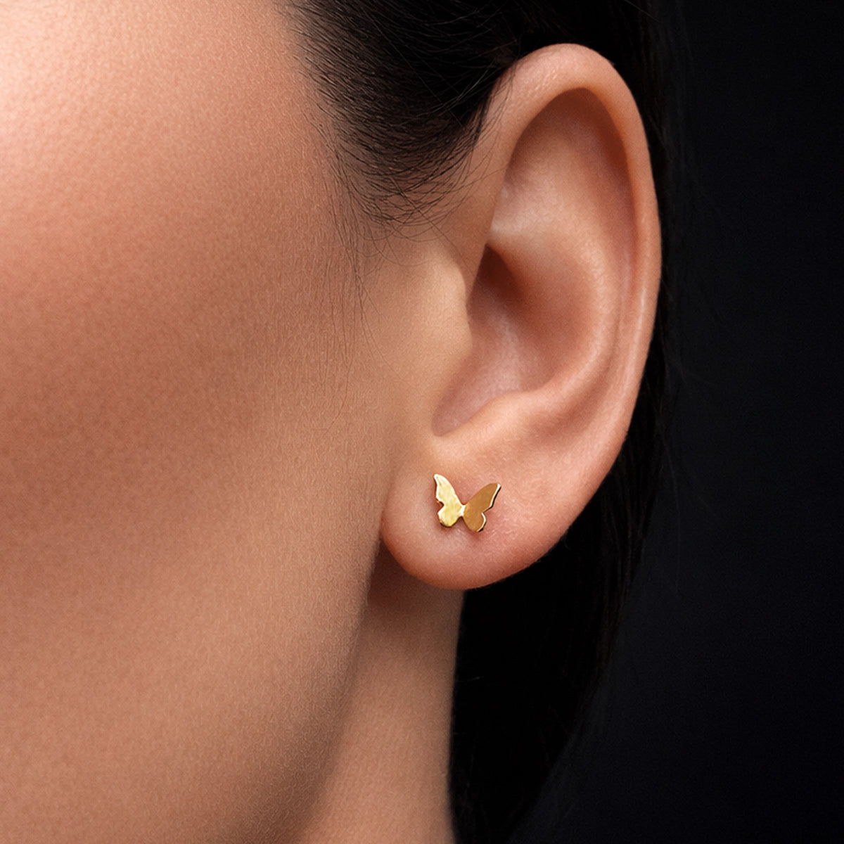Gold butterfly stud earring in polished 18k gold tone, worn on a model’s ear, minimalist dainty jewelry, small butterfly post earring, delicate everyday accessory, close-up studio shot with smooth skin and dark background.