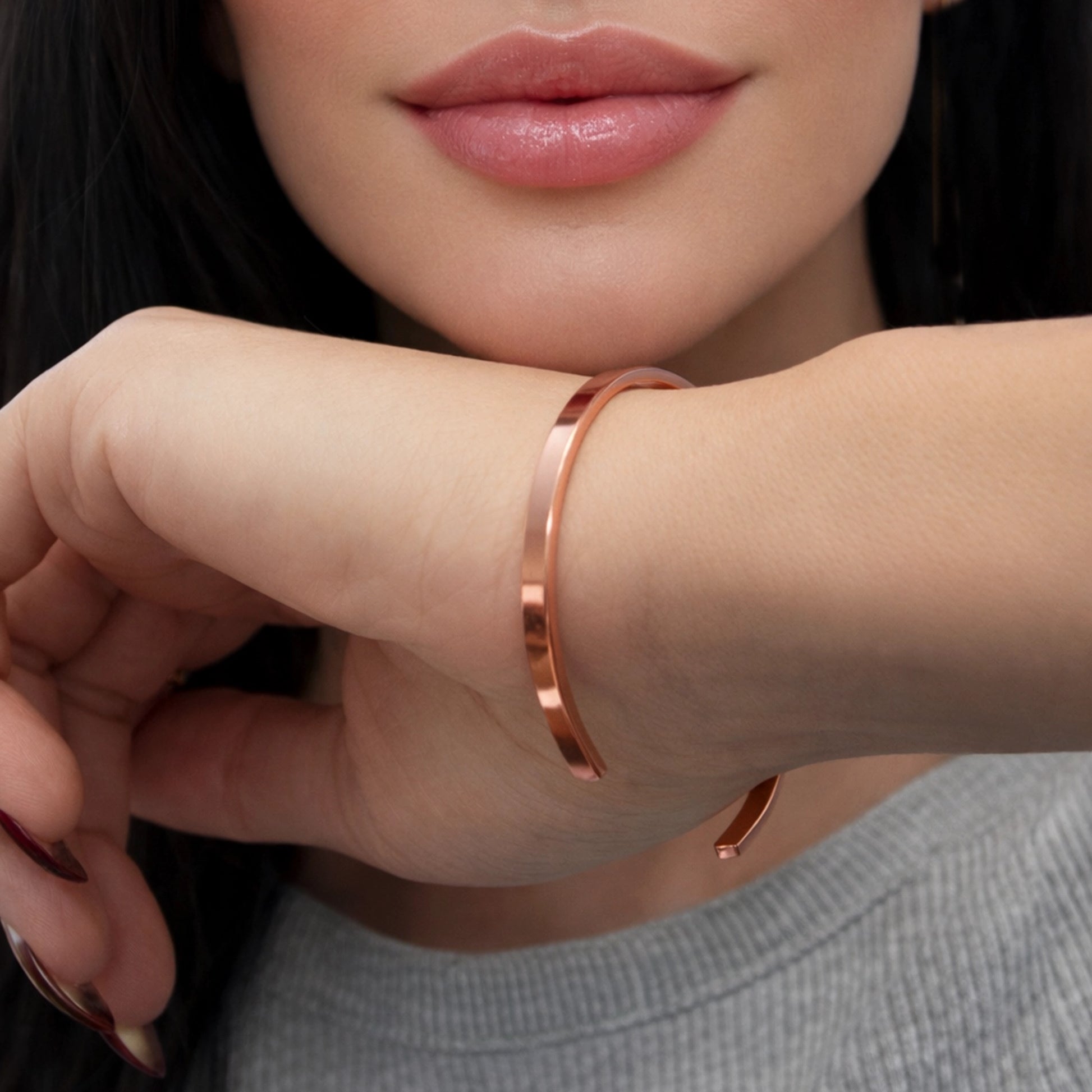 Close-up portrait of a beautiful woman with sensual lips and flawless skin, resting her hand near her face while wearing a minimalist open copper cuff bracelet. The natural copper tone is clearly visible, not gold, with a smooth polished finish. Elegant studio lighting, square composition, modern jewelry style.