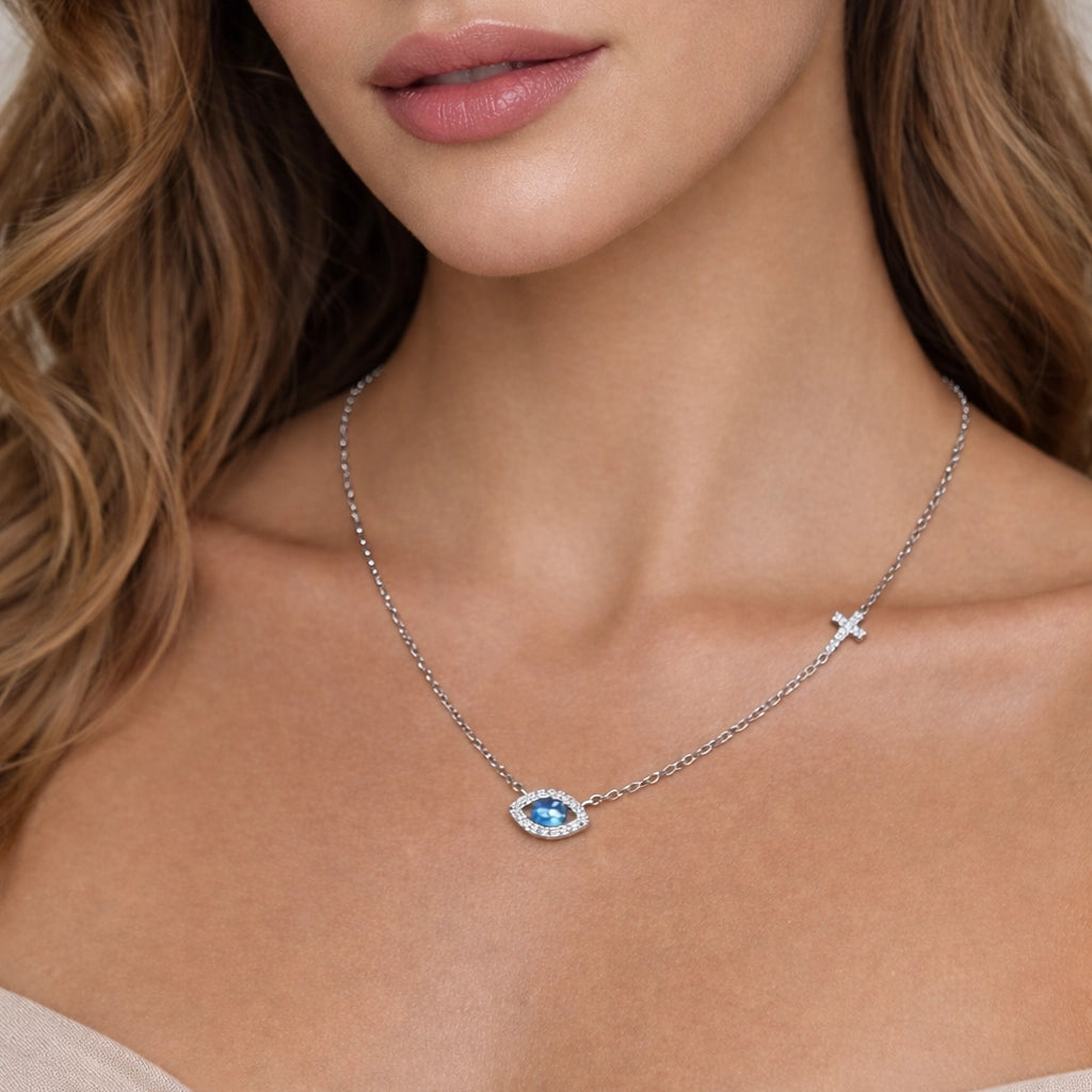 Close-up of a woman wearing a delicate sterling silver necklace featuring a centered blue evil eye charm framed in white cubic zirconia, paired with a small sideways cross charm on the chain, symbolizing protection and faith.
