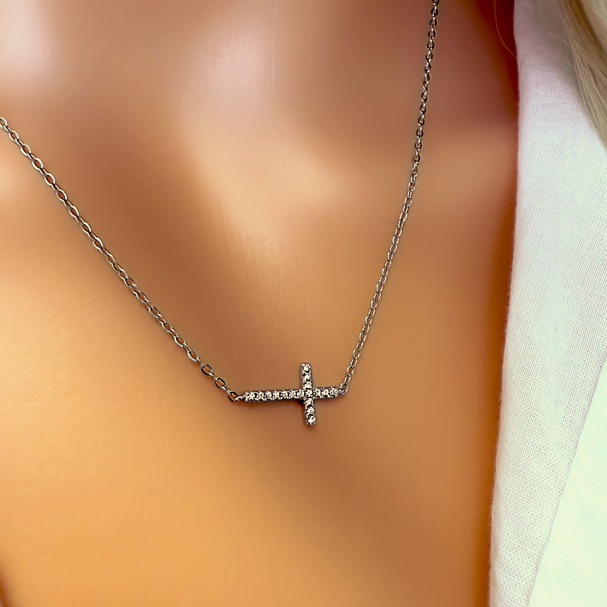 Macro view of a silver sideways cross pendant necklace on a mannequin’s neckline, emphasizing the sparkling cubic zirconia details against smooth skin and a soft white shirt.