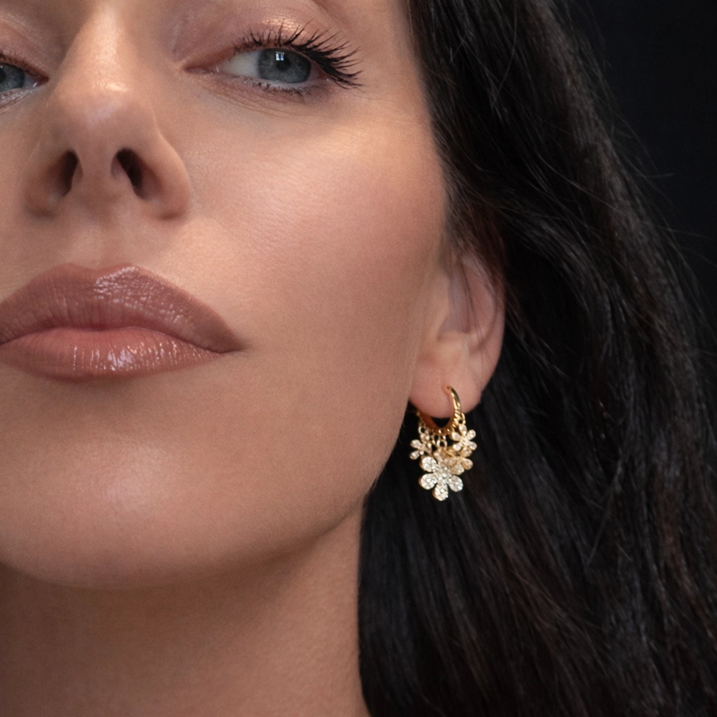 Dainty gold floral drop earrings with sparkling cubic zirconia worn on a beautiful model, close-up professional studio jewelry photo highlighting delicate flower charms, gold huggie hoop earrings, elegant minimalist gold jewelry, feminine luxury earring detail, high-end jewelry photography, modern gold flower earrings, bridal and everyday fine jewelry, stylish gold earrings on model, premium gold filled floral earrings.