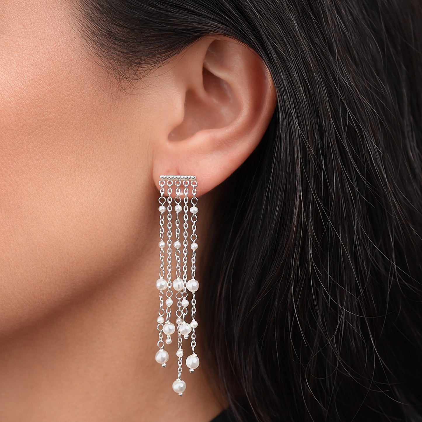Silver pearl chain fringe earrings on model, featuring a horizontal textured bar stud with multiple delicate dangling chains adorned with small white pearls, elegant cascading design, close-up side profile shot with smooth skin and dark hair, soft natural lighting.
