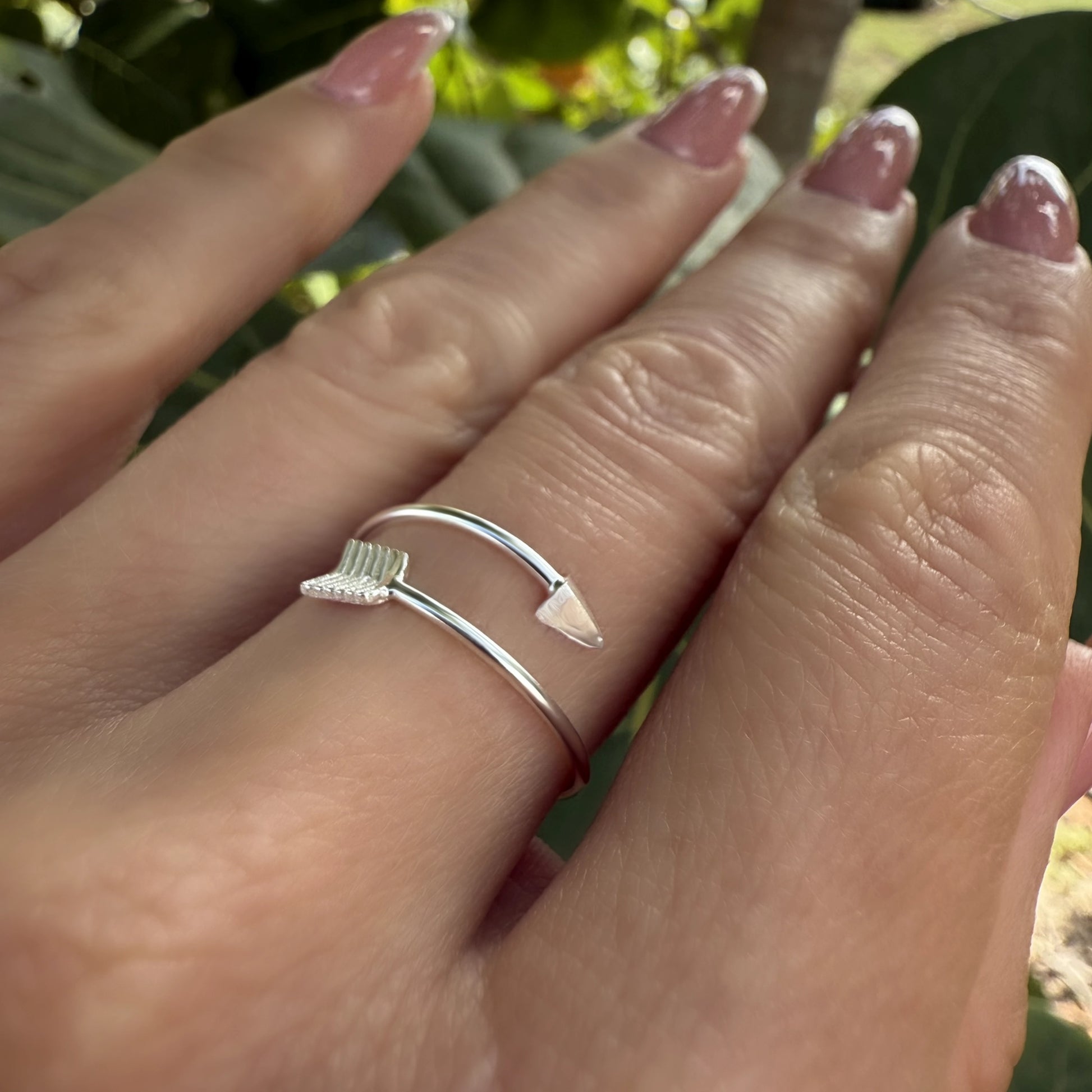 Hand wearing a minimalist sterling silver arrow wrap ring with an open design, photographed outdoors against green leaves and natural sunlight.