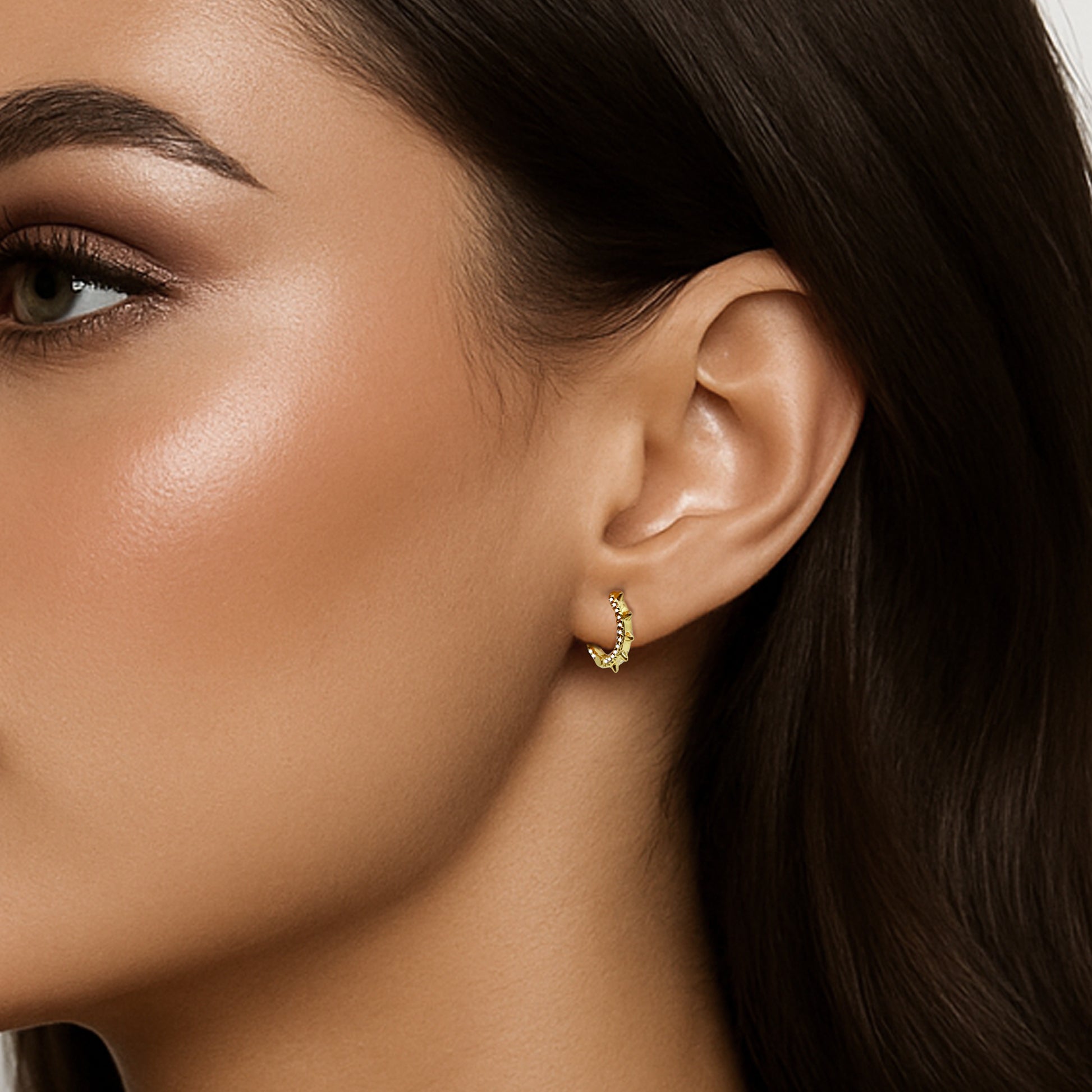 Close-up side view of a woman wearing a small 18k gold filled spike hoop earring with cubic zirconia accents, styled on her earlobe with smooth, polished makeup and dark brown hair.
