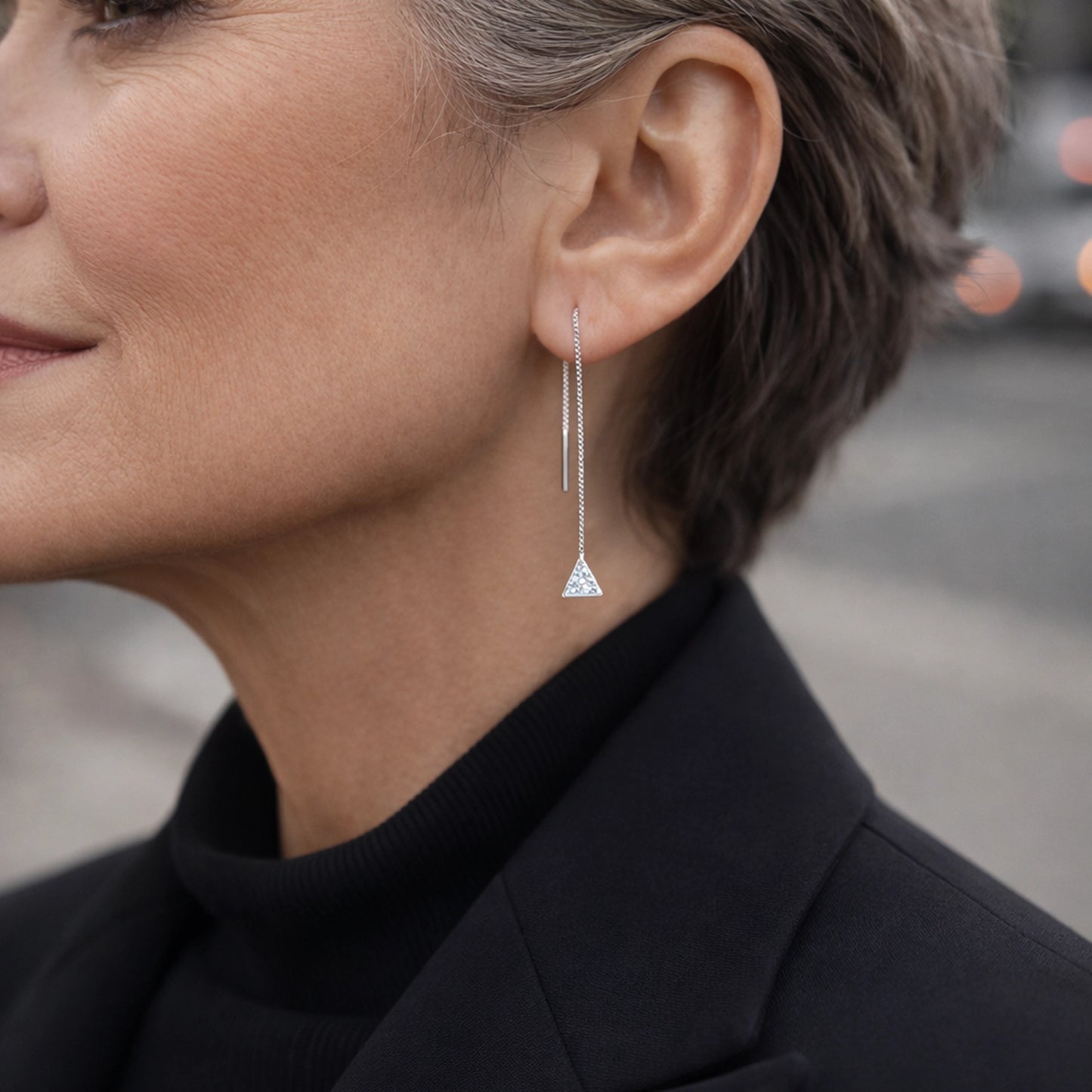 Modern sterling silver threader earrings with double chain and small triangle CZ pendant, worn by stylish mature woman in black blazer, elegant minimalist dangle earrings photographed in soft natural city light.