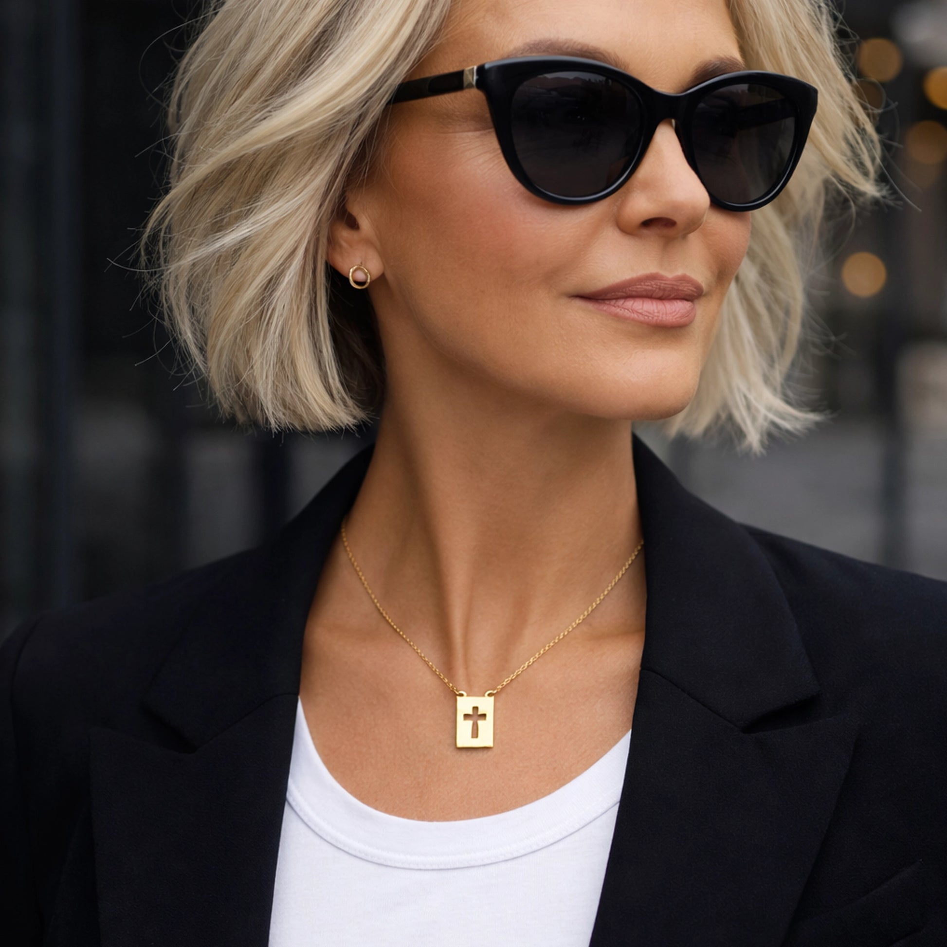Elegant mature woman wearing gold cross pendant necklace on delicate chain, stylish modern outfit with black blazer and white top, professional studio lighting, sharp focus, luxury jewelry portrait, sophisticated fashion model in her 50s, gold religious necklace close-up, contemporary fashion background.
