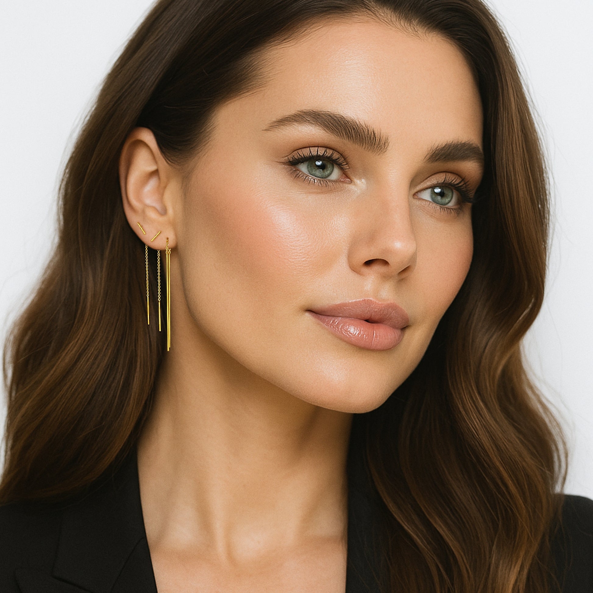 Close-up portrait of a beautiful woman with glowing skin and wavy brown hair wearing elegant gold bar threader chain earrings. The earrings feature delicate chains with sleek vertical bars, styled against her ear in a modern layered look. The model wears a black blazer, and the background is clean white, emphasizing the jewelry.