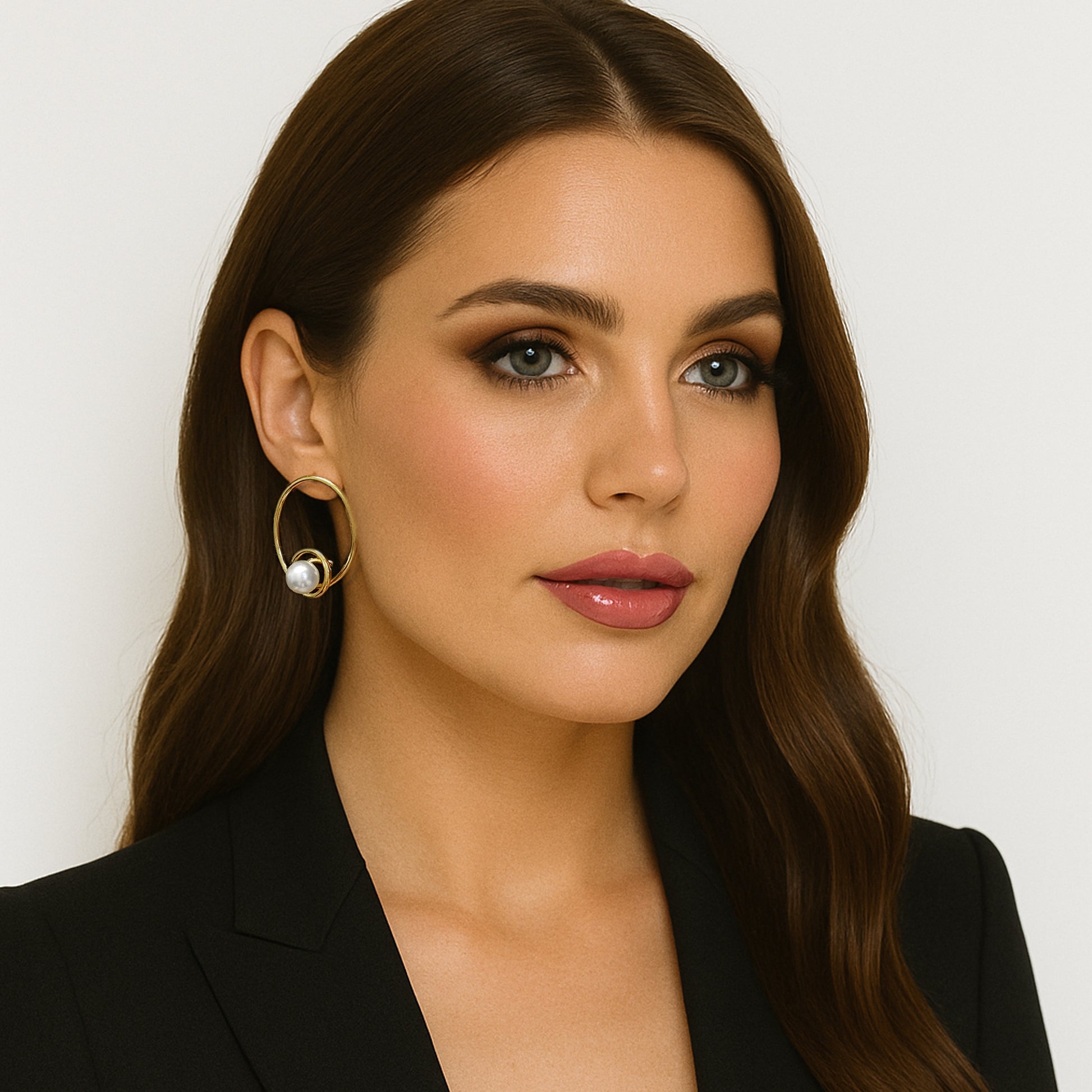 Close-up of a woman with long brown hair and flawless makeup wearing elegant gold hoop earrings. Each hoop features a large round pearl nestled inside the curve of the hoop. She is dressed in a black blazer, and the clean white background highlights the sophisticated design of the earrings.