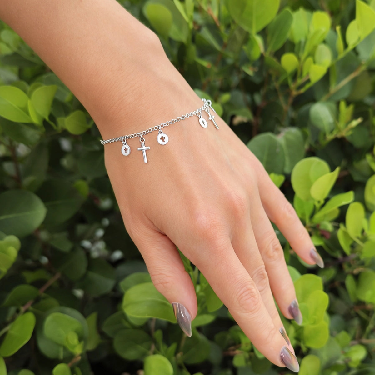 Sterling silver religious charm bracelet on hand with cross and medallion dangles, displayed outdoors against vibrant green foliage.