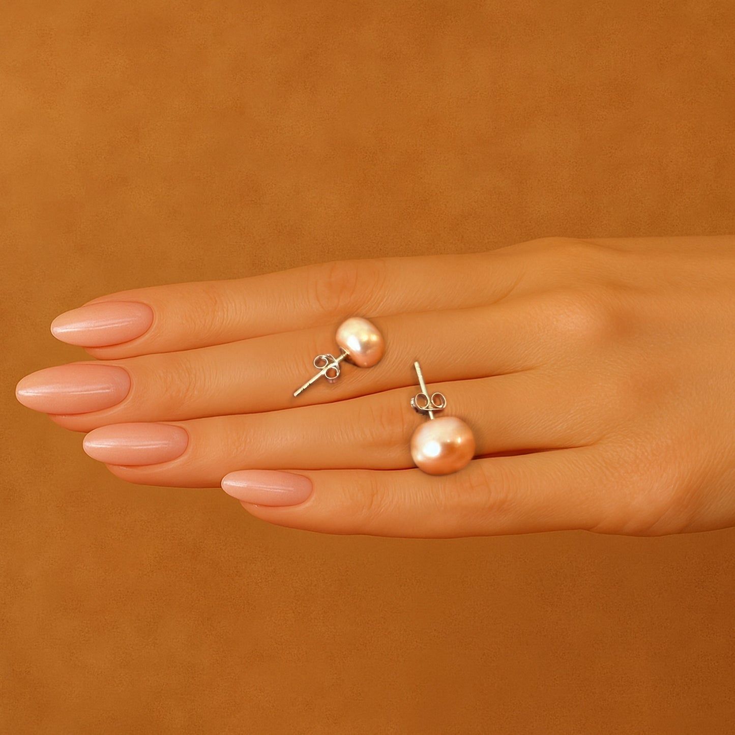 Real peach freshwater pearl stud earrings displayed on a hand with manicured nails, highlighting their natural glow and sterling silver push-back posts.