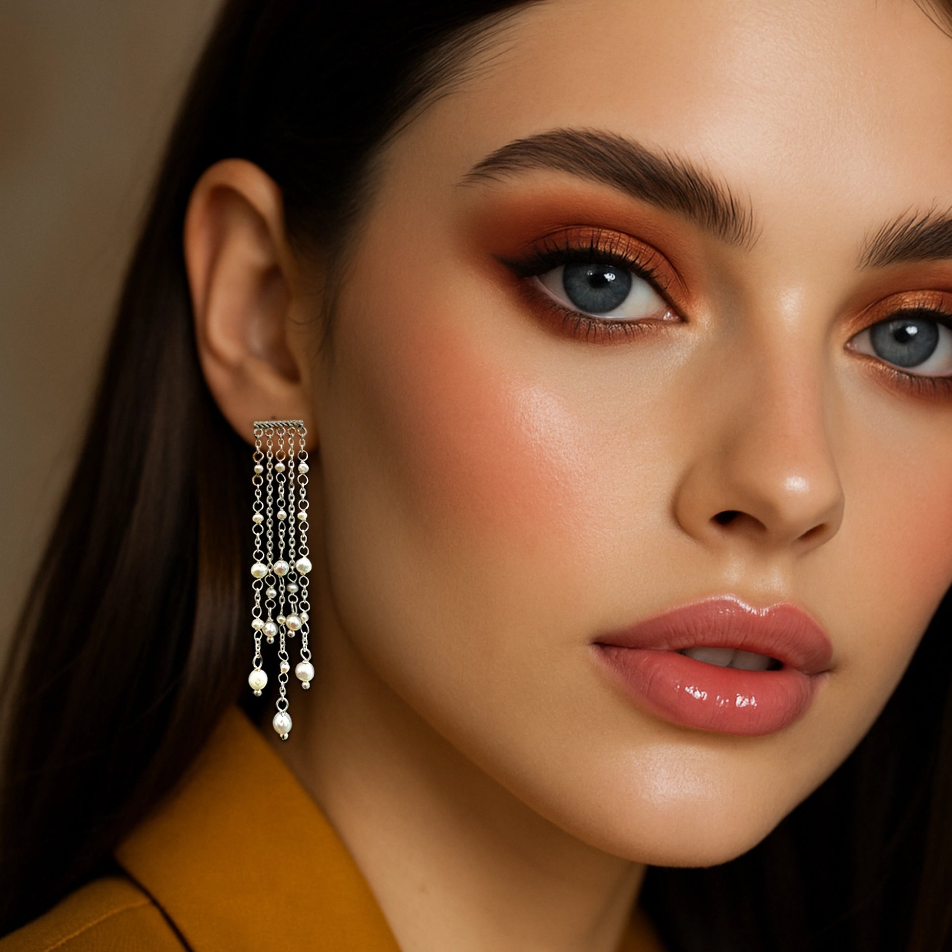 Close-up of a woman wearing long chandelier drop earrings with natural freshwater pearls and silver chain strands, styled with glossy lips and a mustard blazer.