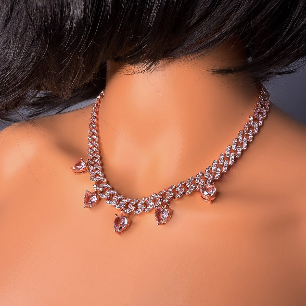 Close-up of a rose gold curb chain necklace with sparkling cubic zirconia and dangling pink heart-shaped charms on a mannequin with short dark hair.