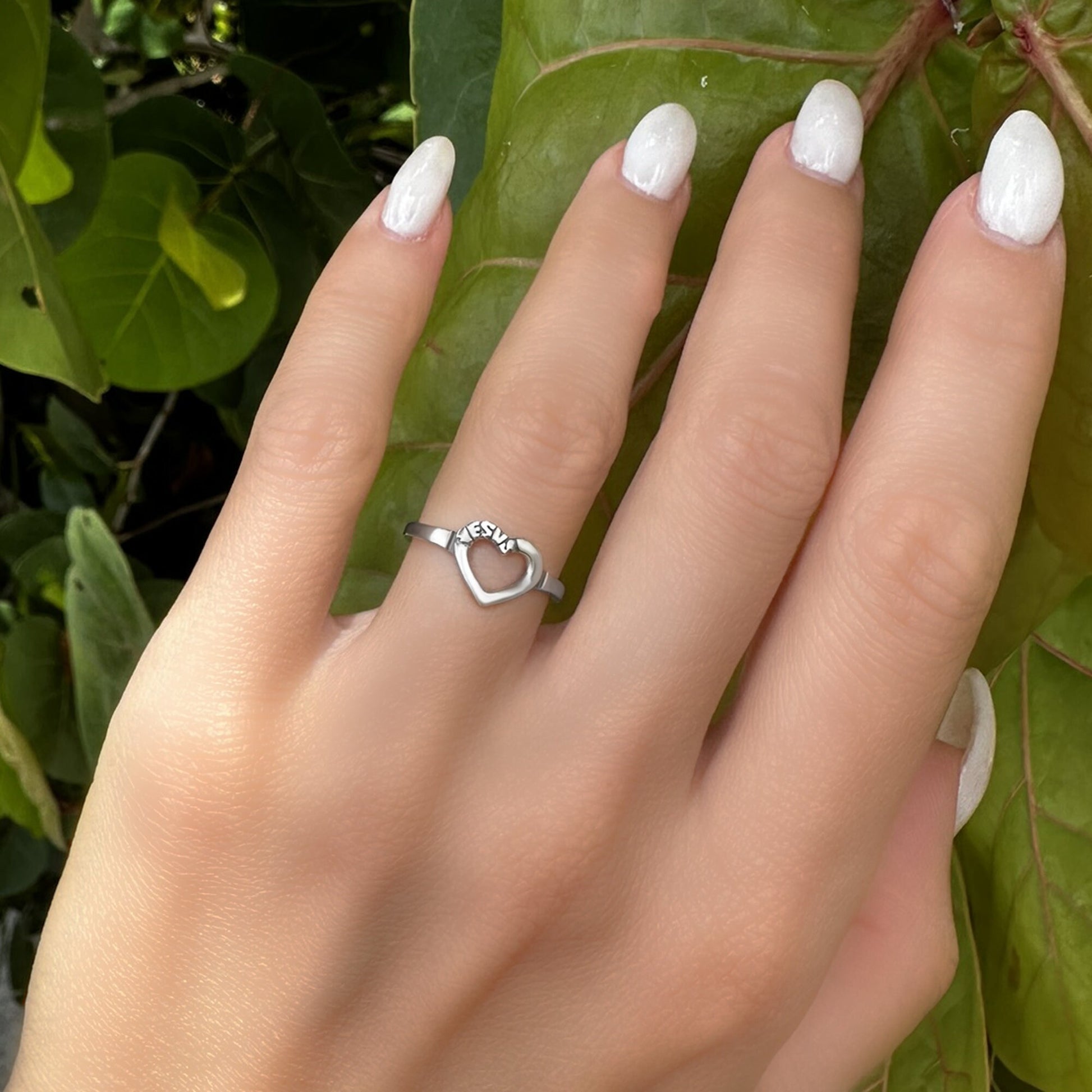 A sterling silver ring featuring an open heart engraved with “Jesus,” worn on a hand with white nails against a leafy green background.