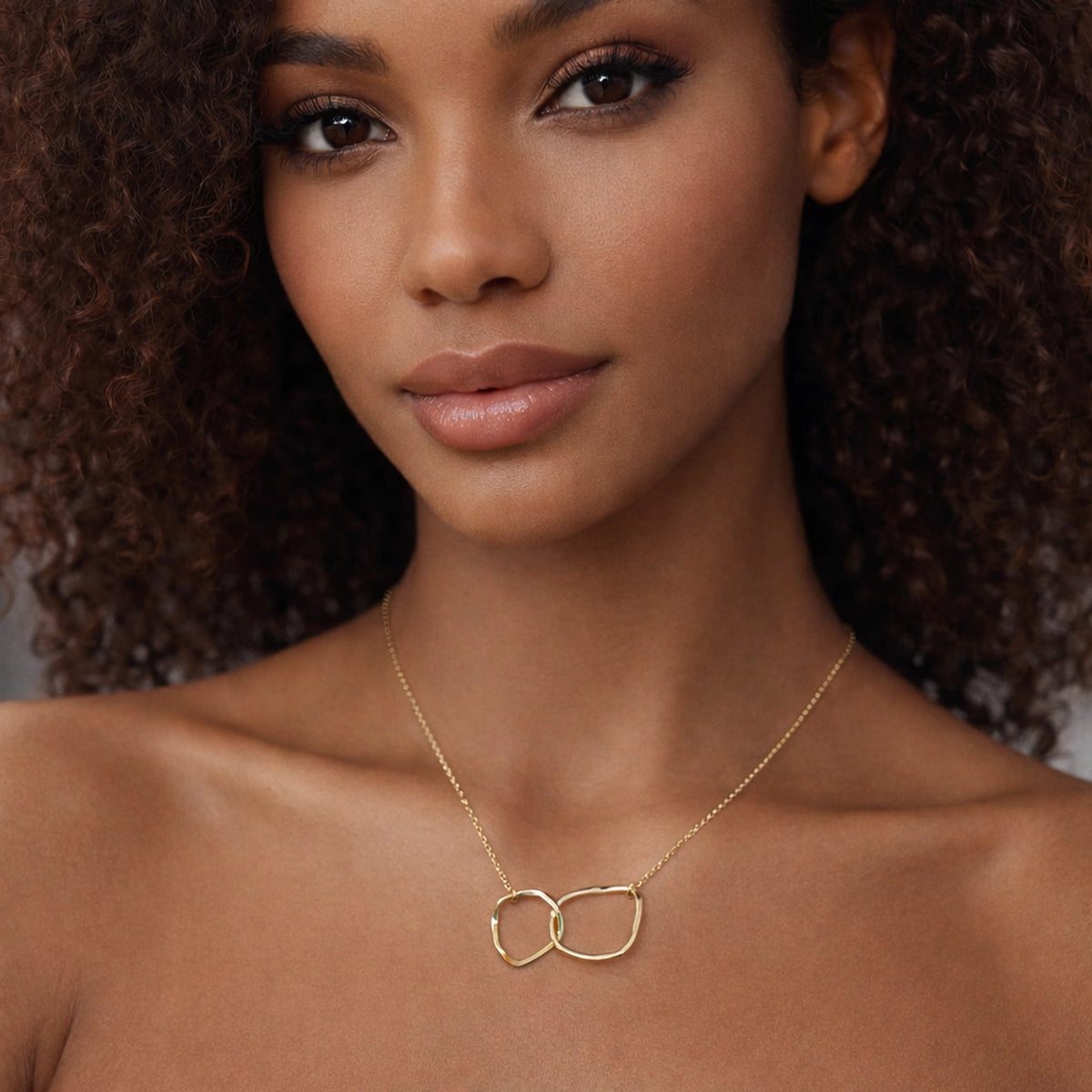 beautiful  model wearing a delicate gold chain necklace with two interlocking organic-shaped gold charms, shown in sharp focus with professional lighting and a soft neutral background.