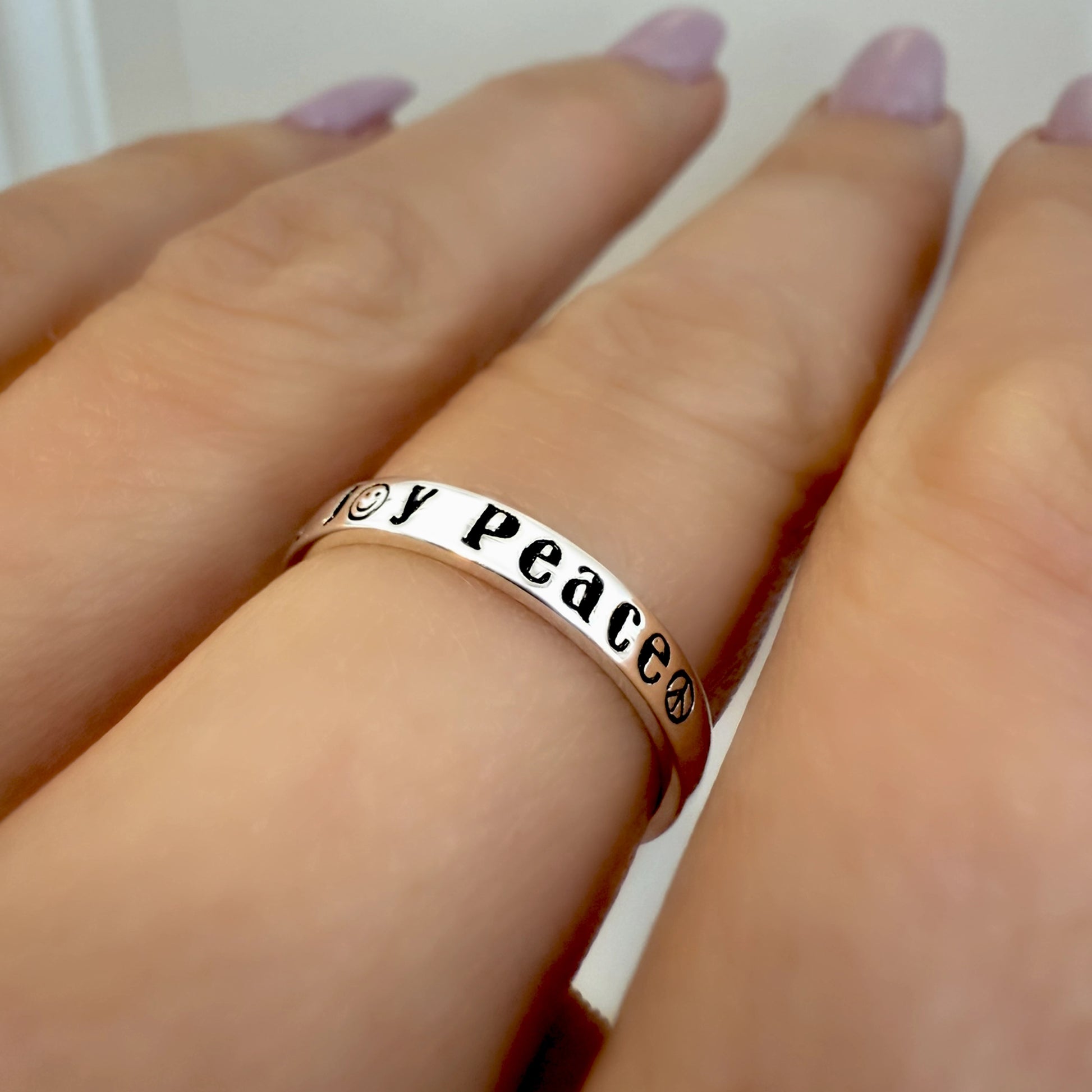 Sterling silver ring engraved with the words "Joy" and "Peace" featuring a smiley face and peace symbol, worn on a hand with light pink nails.