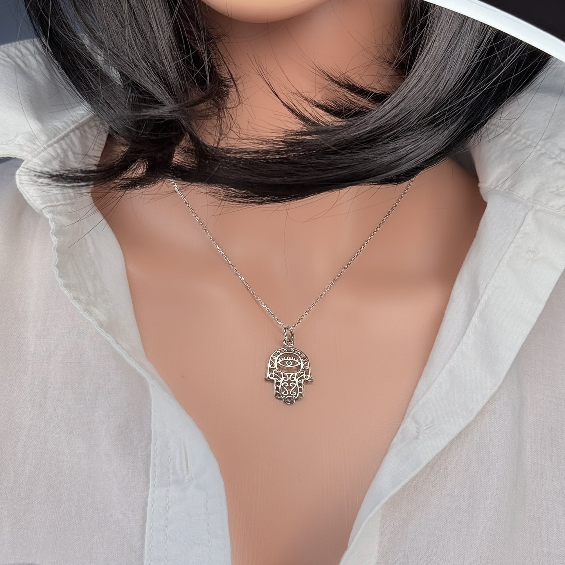 Close-up of a mannequin wearing a sterling silver Hamsa hand pendant necklace with an intricate lace design and evil eye detail, styled with an open-collar white shirt.