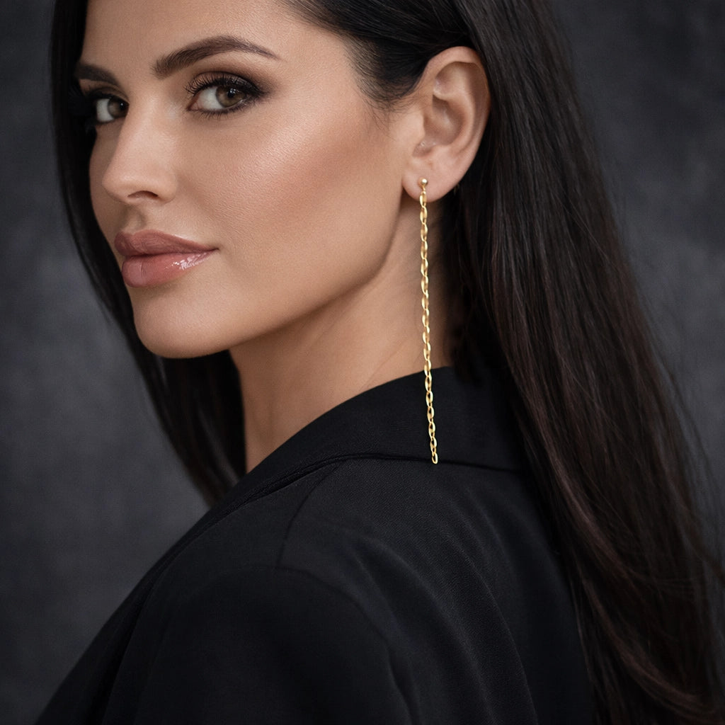 lose-up portrait of a woman in a black oversized blazer, turned in three-quarter profile, wearing a single long gold chain drop earring. Flawless even skin tone, glossy lips, sleek dark hair, professional studio lighting with a soft blurred background, sharp focus on the earring.
