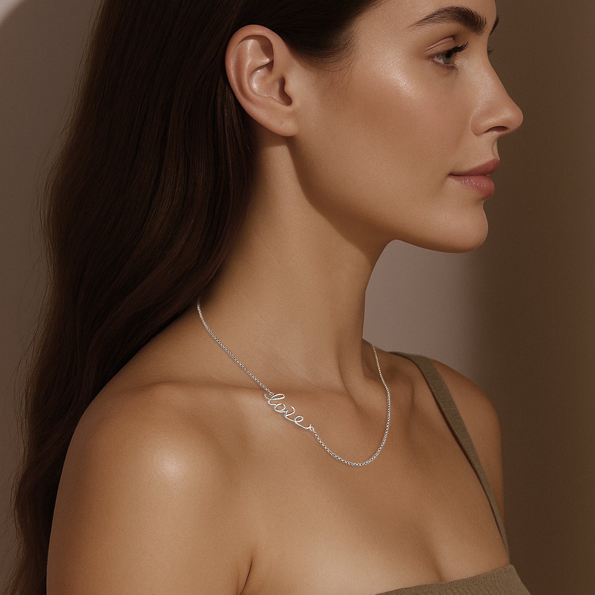 A woman with long brown hair and glowing skin wearing a sterling silver script necklace that spells ‘love.’ She is shown in side profile with a neutral-toned background, wearing a beige top.