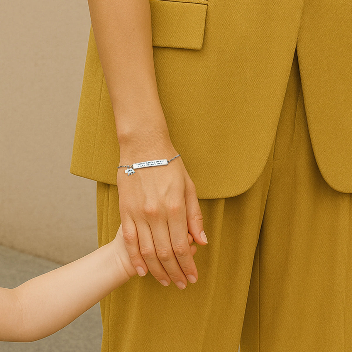Sterling silver bracelet engraved with the message "There is nothing greater than a mother's love", featuring a tiny baby bear charm. A woman in a mustard suit lovingly holds a child's hand, showcasing the meaningful gift.