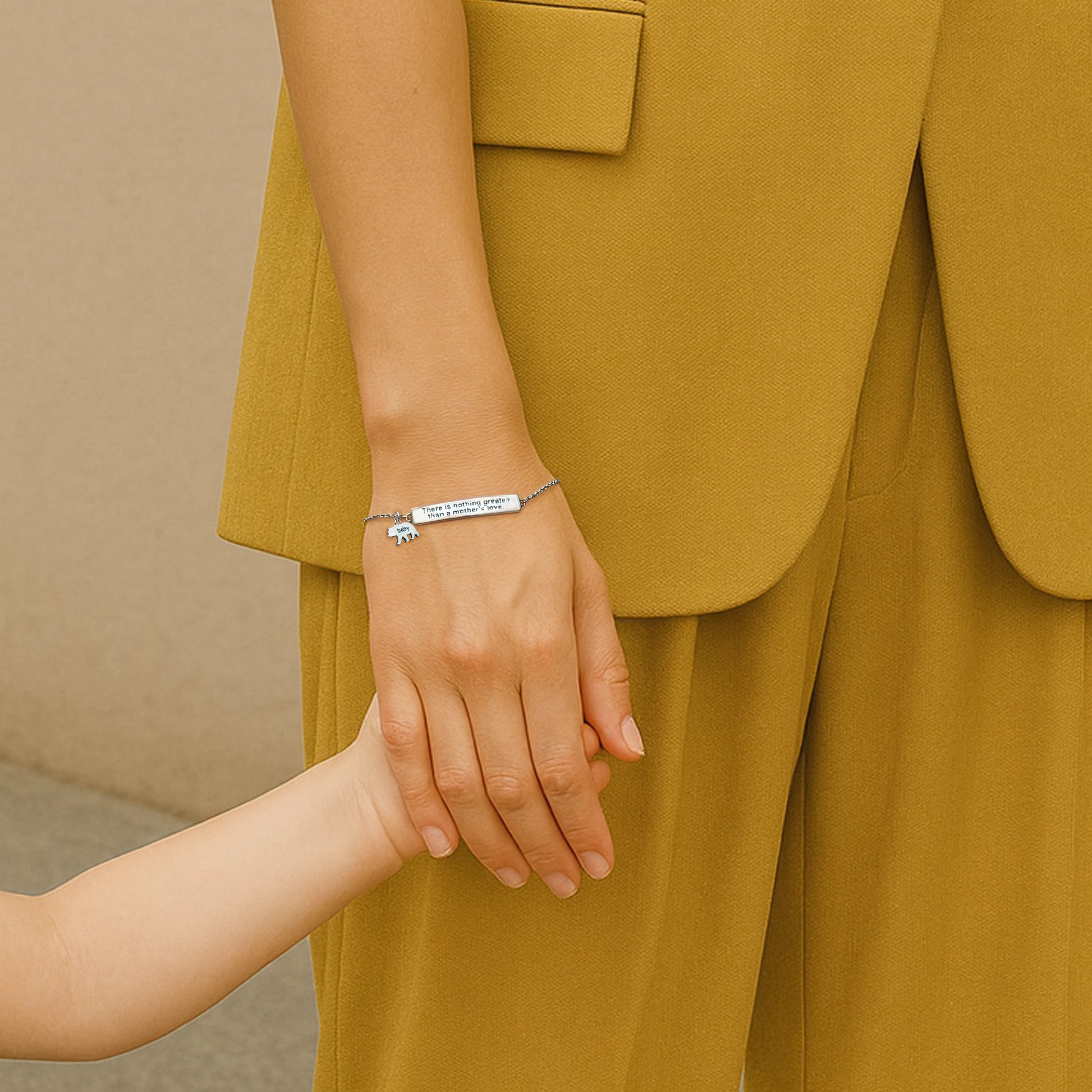 Sterling silver bracelet engraved with the message "There is nothing greater than a mother's love", featuring a tiny baby bear charm. A woman in a mustard suit lovingly holds a child's hand, showcasing the meaningful gift.