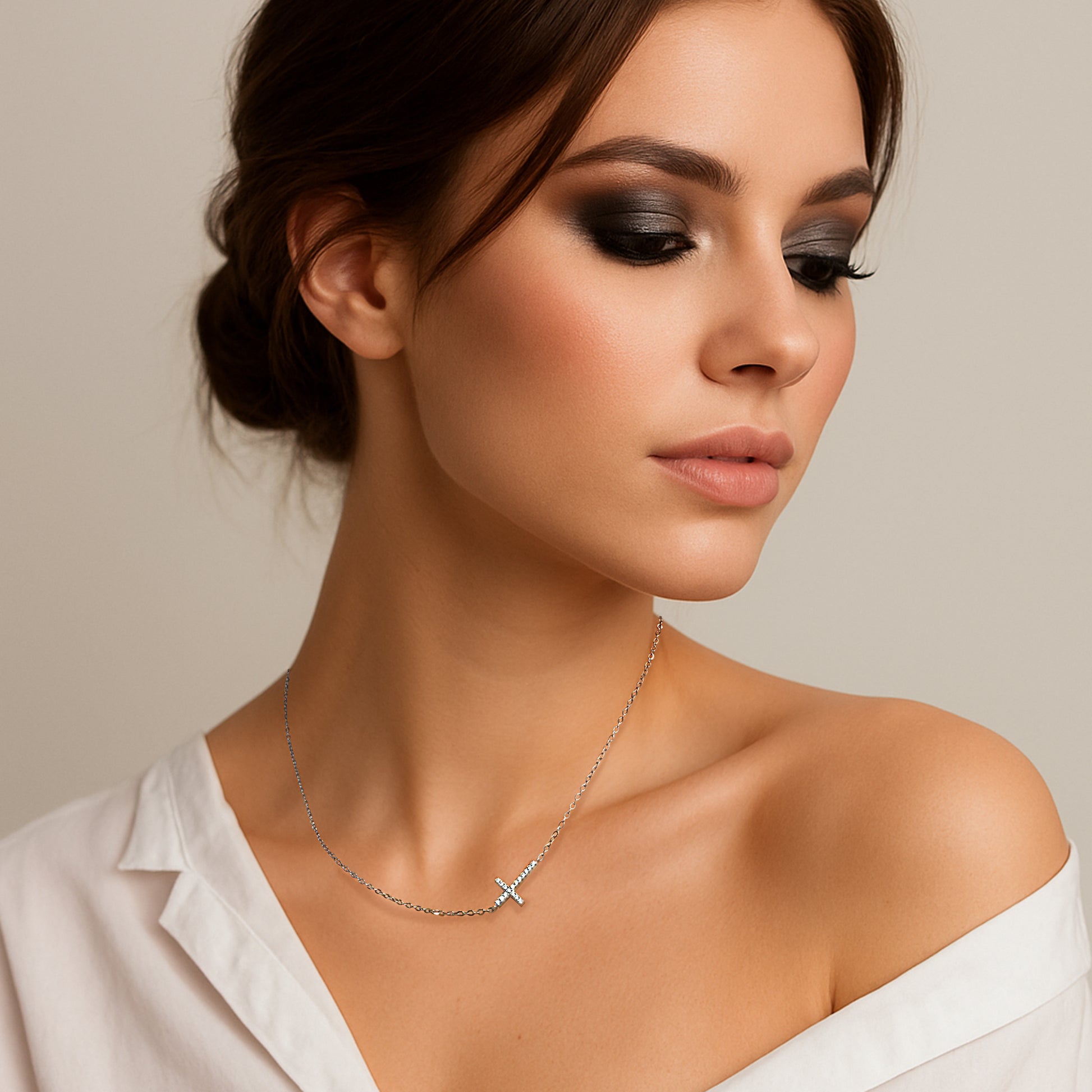 Beautiful model with smoky eye makeup wearing a minimalist silver sideways cross necklace; her white shirt elegantly falls off one shoulder, highlighting a soft and elegant neckline.