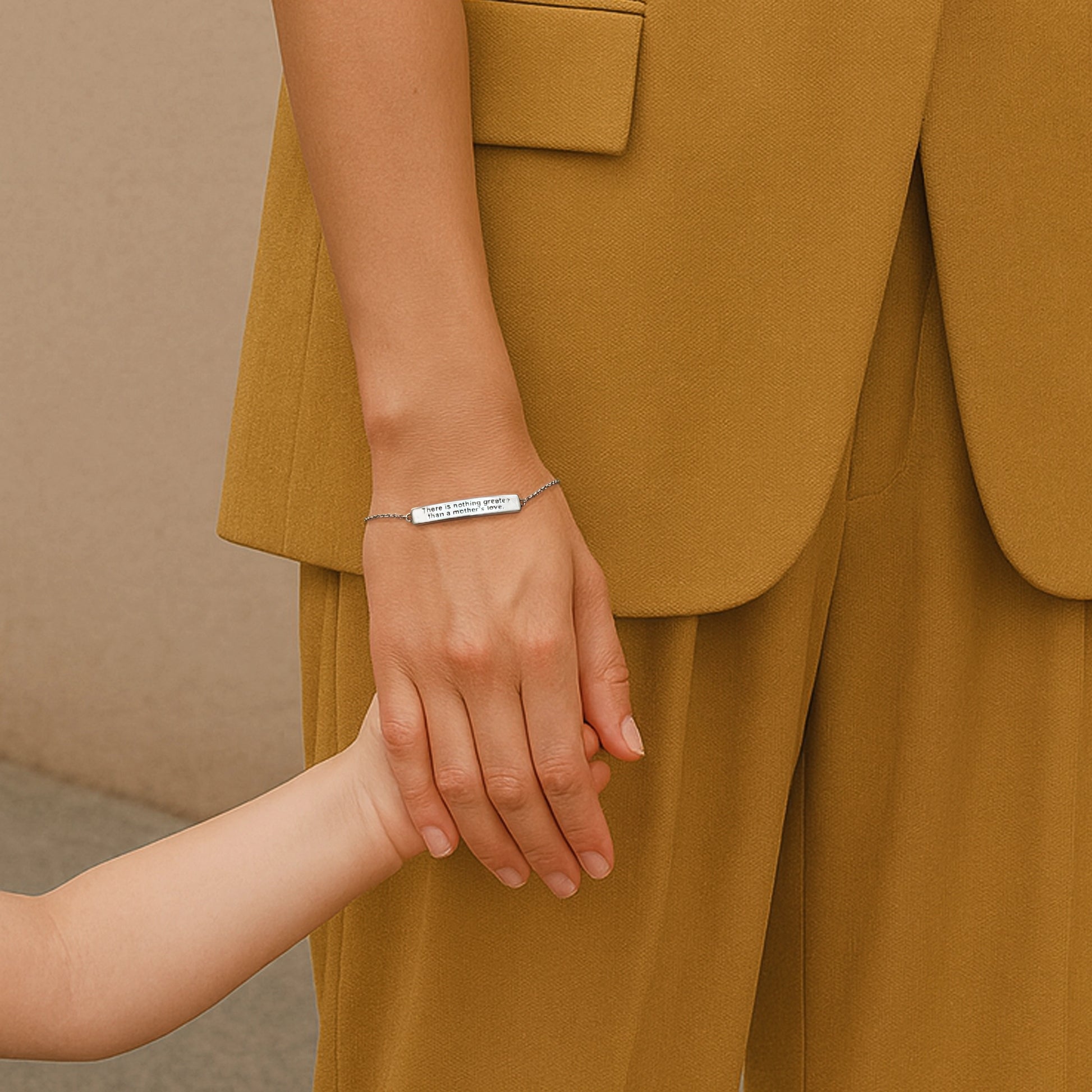 A woman in a mustard-colored suit holding a child’s hand, wearing a sterling silver engraved bar bracelet that reads “There is nothing greater than a mother’s love.”