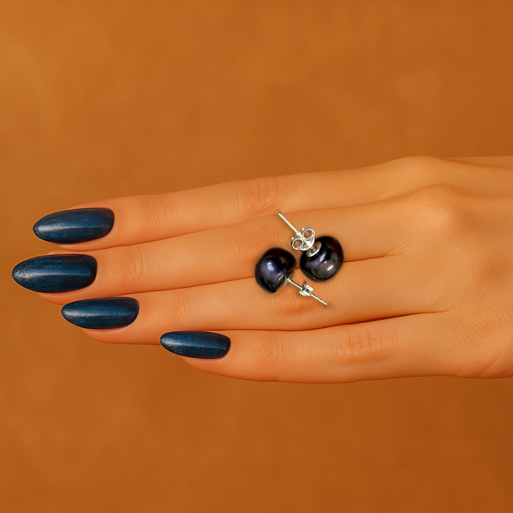 A pair of dark blue freshwater pearl stud earrings with sterling silver posts resting on a woman’s hand, styled with glossy navy blue manicure against a warm brown background.