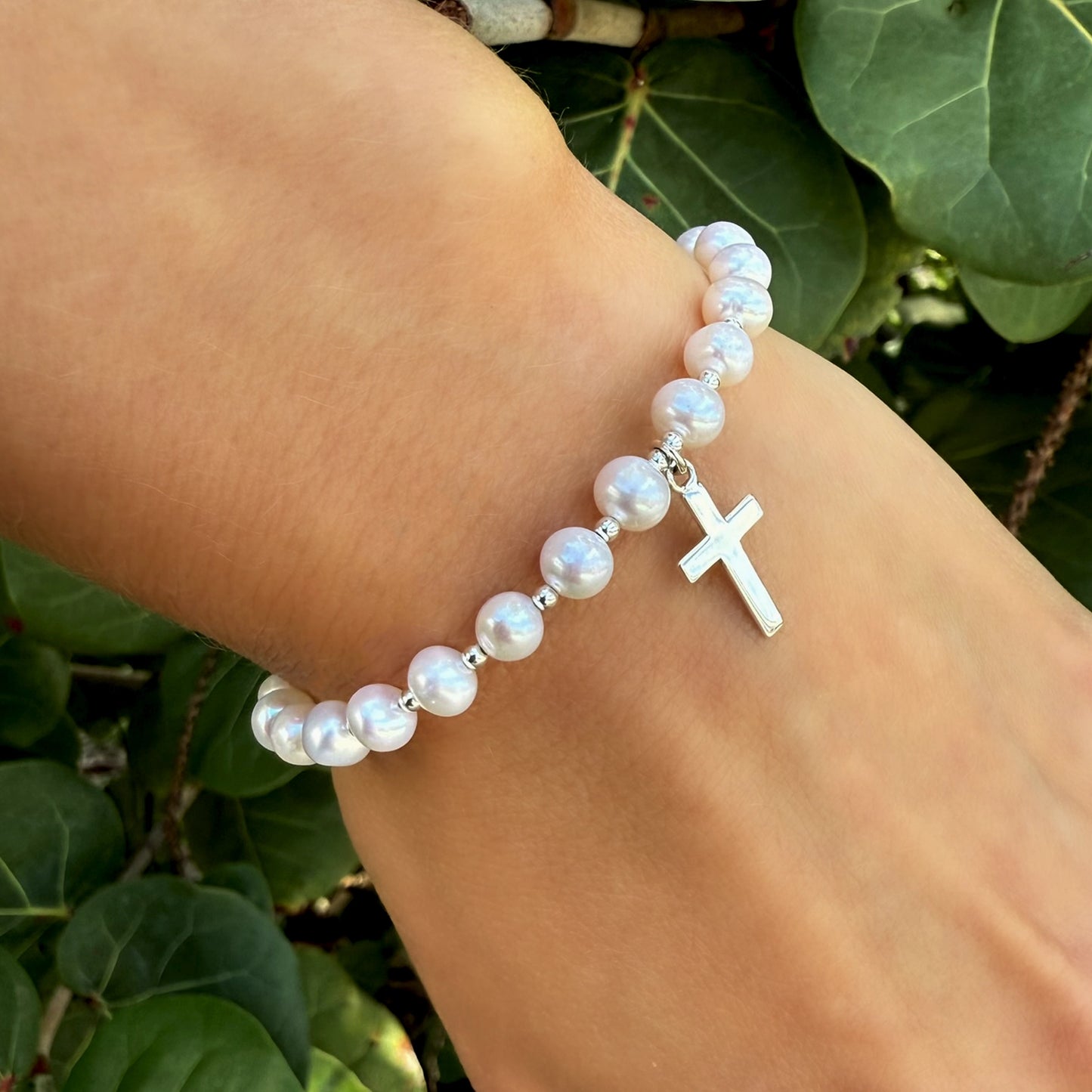 Natural white freshwater pearl bracelet featuring a dangling silver cross charm, shown on a wrist surrounded by lush green leaves.