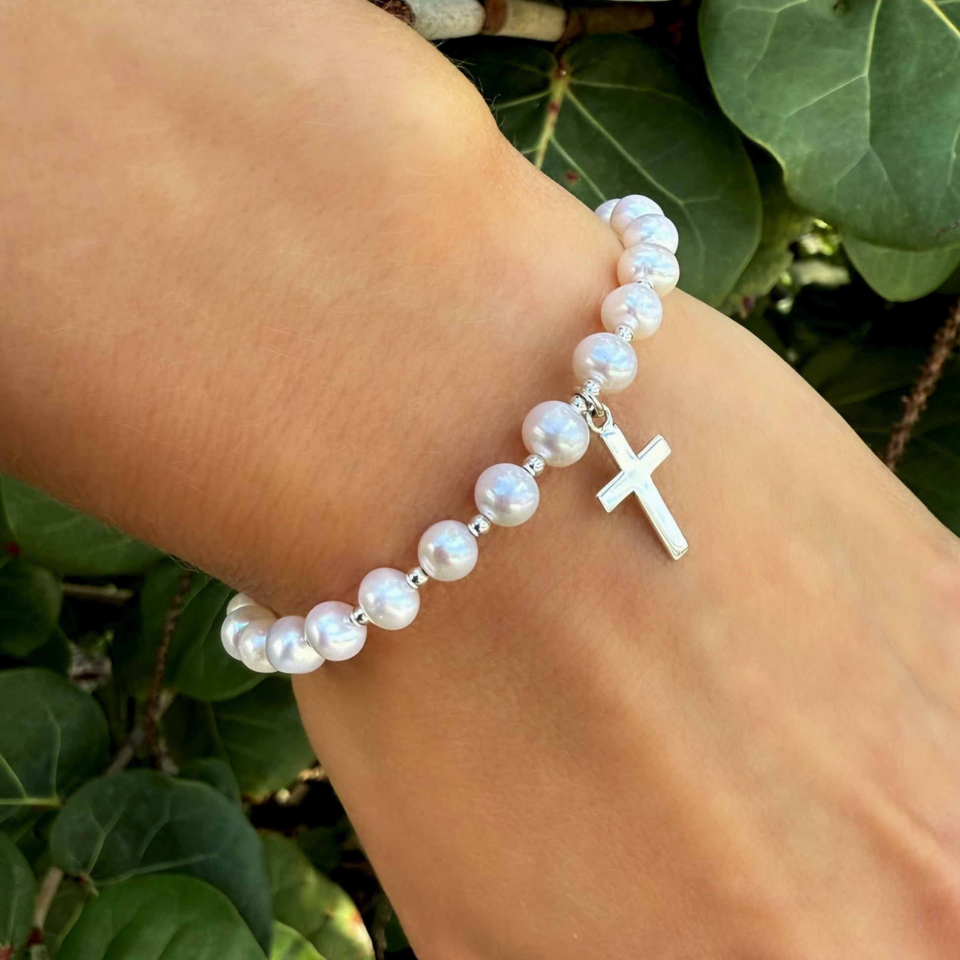 Natural white freshwater pearl bracelet featuring a dangling silver cross charm, shown on a wrist surrounded by lush green leaves.