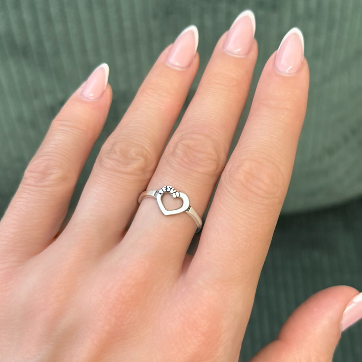 A woman's hand with soft pink almond-shaped nails wearing a sterling silver ring featuring an open heart engraved with the name “Jesus,” set against a muted green textured background.