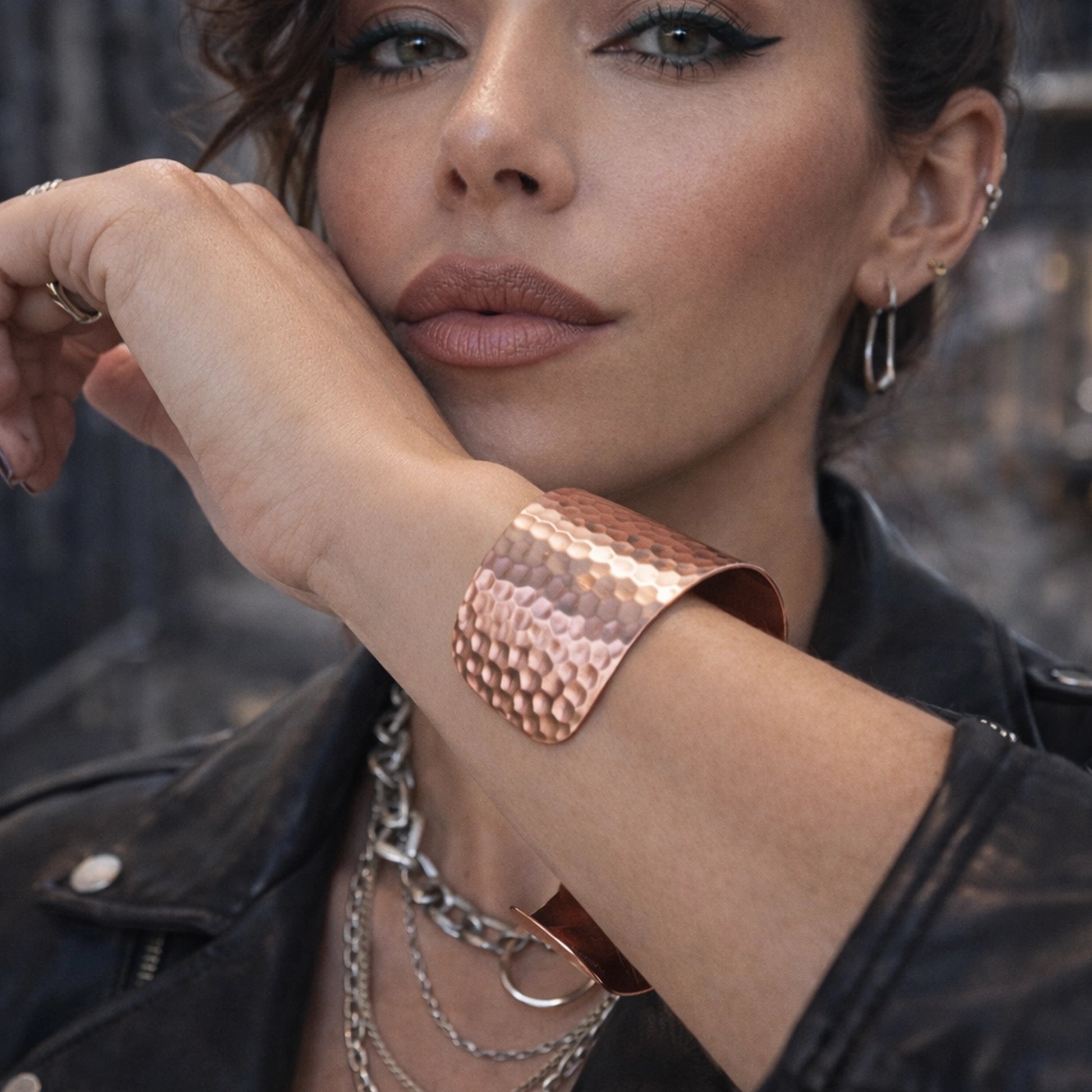 Oversized open hammered copper cuff bracelet on woman’s wrist, bold modern statement jewelry styled with layered chains and leather jacket