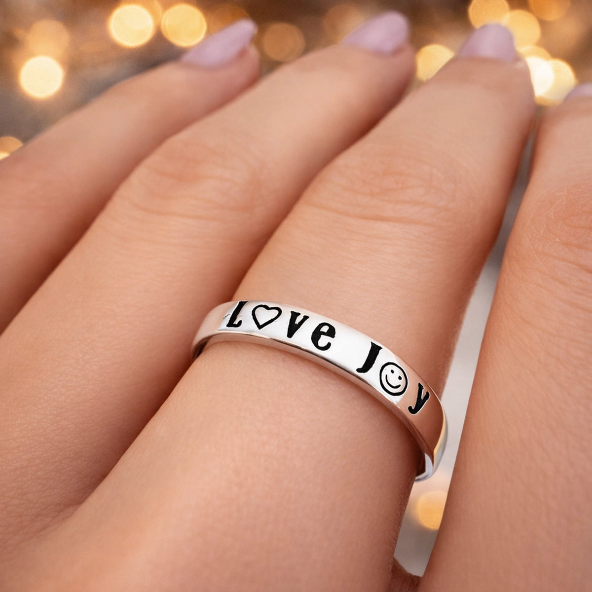 Sterling silver ring engraved with "Love", "Joy", and "Peace" using heart, smiley, and peace symbols, shown on a hand with pastel pink manicure.
