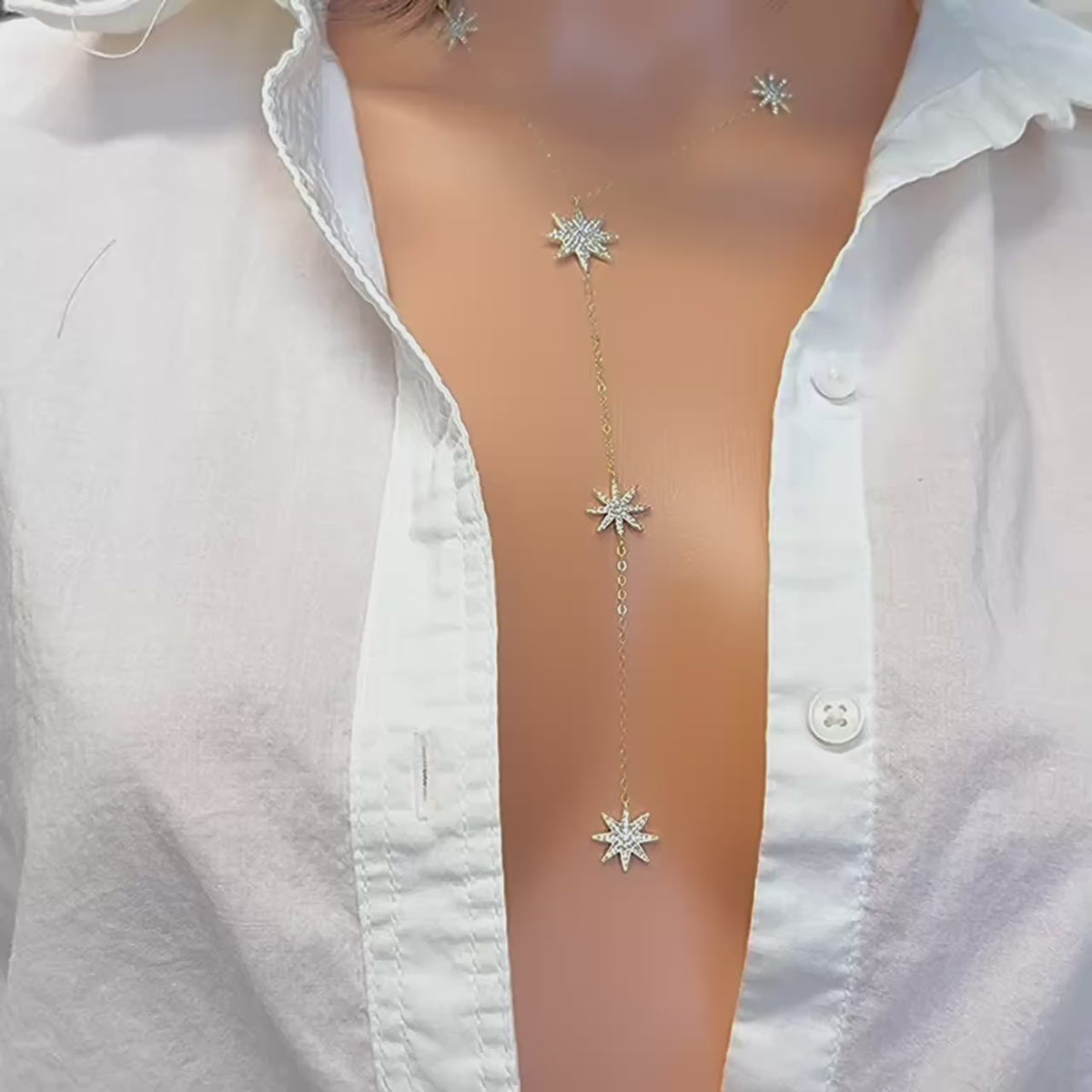 A close-up video of a model wearing a deep V-cut white shirt, showcasing a gold lariat necklace with sparkling CZ star charms cascading down the chest. The delicate chain glimmers in the light as the stars catch movement, creating an elegant and celestial look.