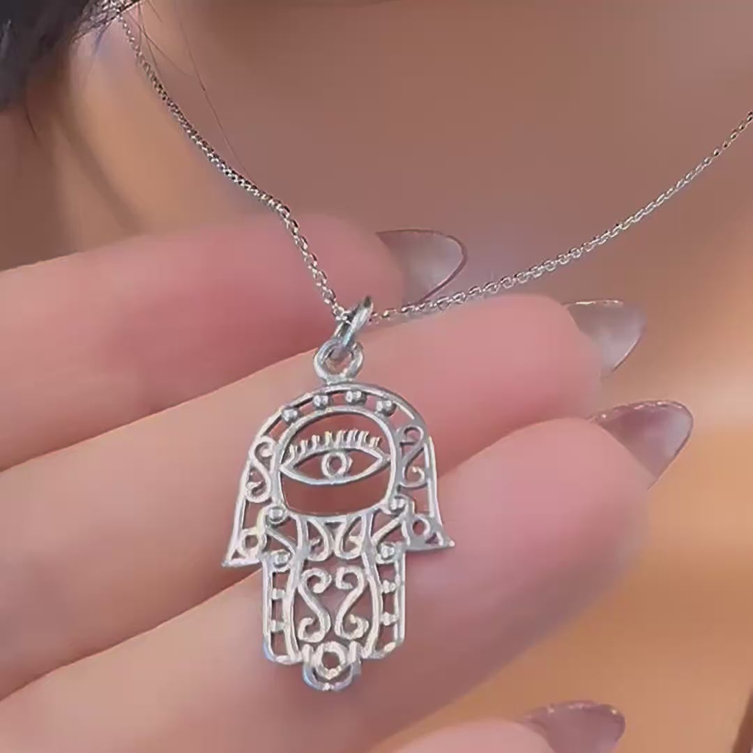 Video of a sterling silver Hamsa hand necklace with intricate lace design and an evil eye detail, displayed on a mannequin wearing an open-collar white shirt.
