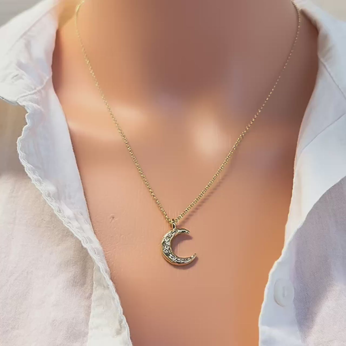 Close-up video of a model wearing a white button-down shirt and a gold-filled necklace featuring a textured crescent moon pendant with sparkling cubic zirconia details. The pendant rests gracefully at the center of the chest, catching the light as it moves.