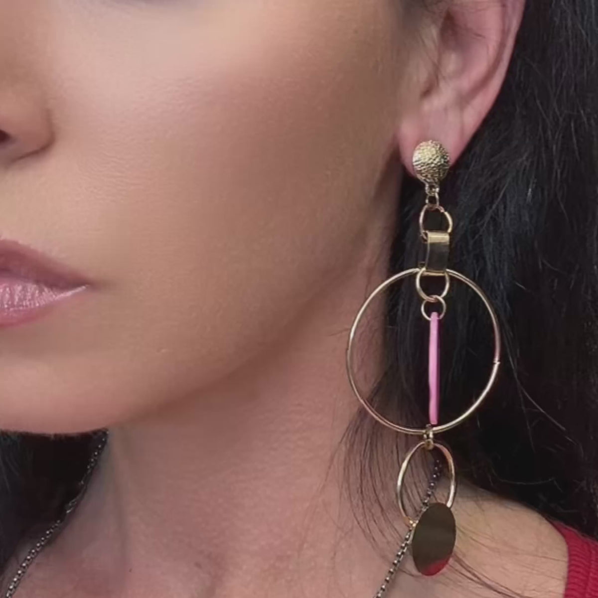Video of an elegant woman showcasing bold asymmetric gold hoop earrings featuring a pink shell disc and hammered metal accents.