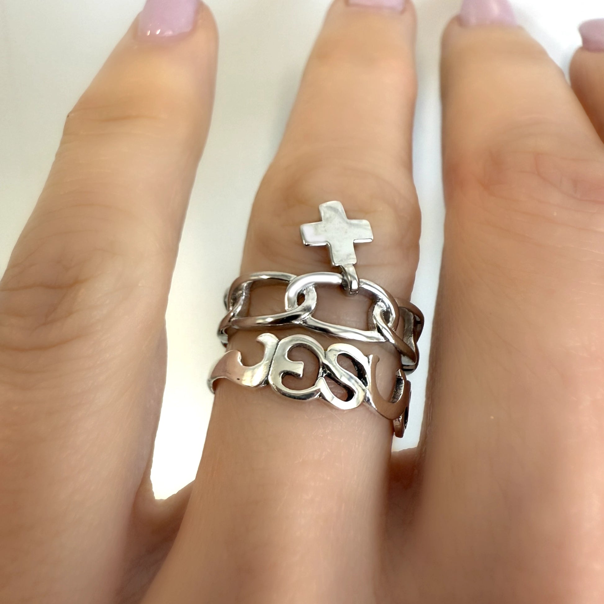 Close-up view of a sterling silver Christian ring set on finger, featuring a “JESUS” cutout band, a chunky chain ring, and a dangling cross charm.