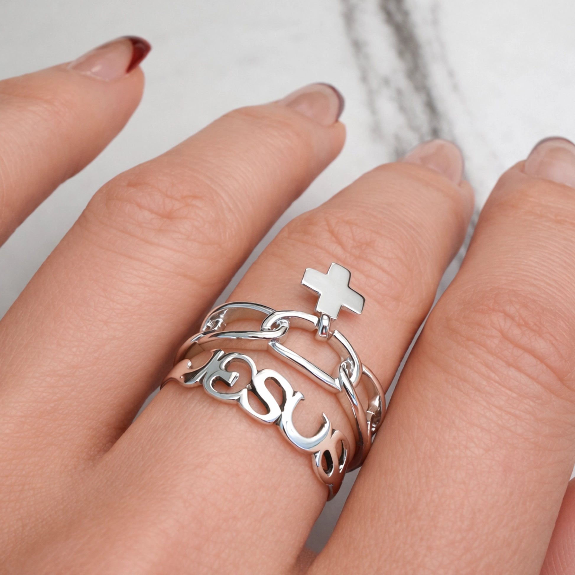Sterling silver Jesus script ring stack with cross charm, shown on a woman walking with hand relaxed at her side, close-up lifestyle jewelry photo.