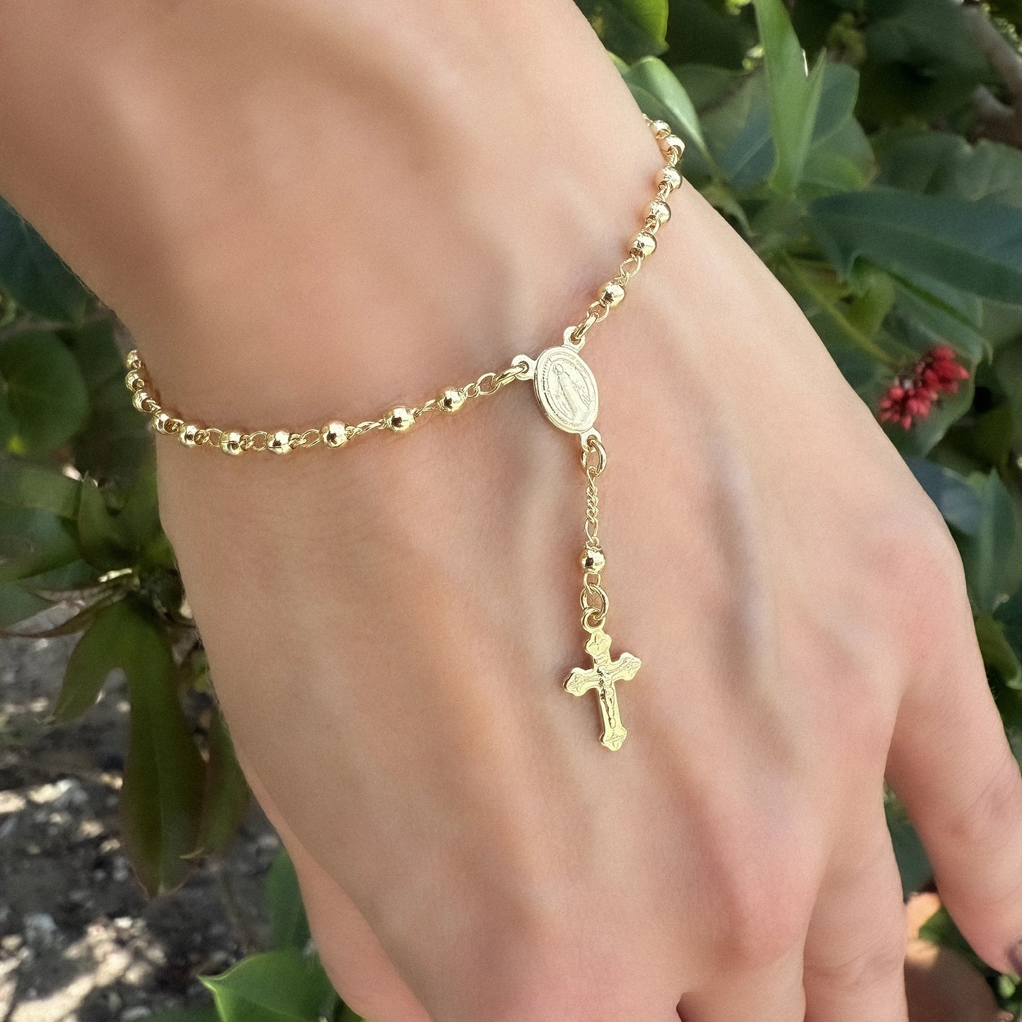 Close-up of a hand wearing a gold filled Virgin Mary rosary bracelet with polished round beads and an ornate dangling cross charm, photographed in natural outdoor lighting.