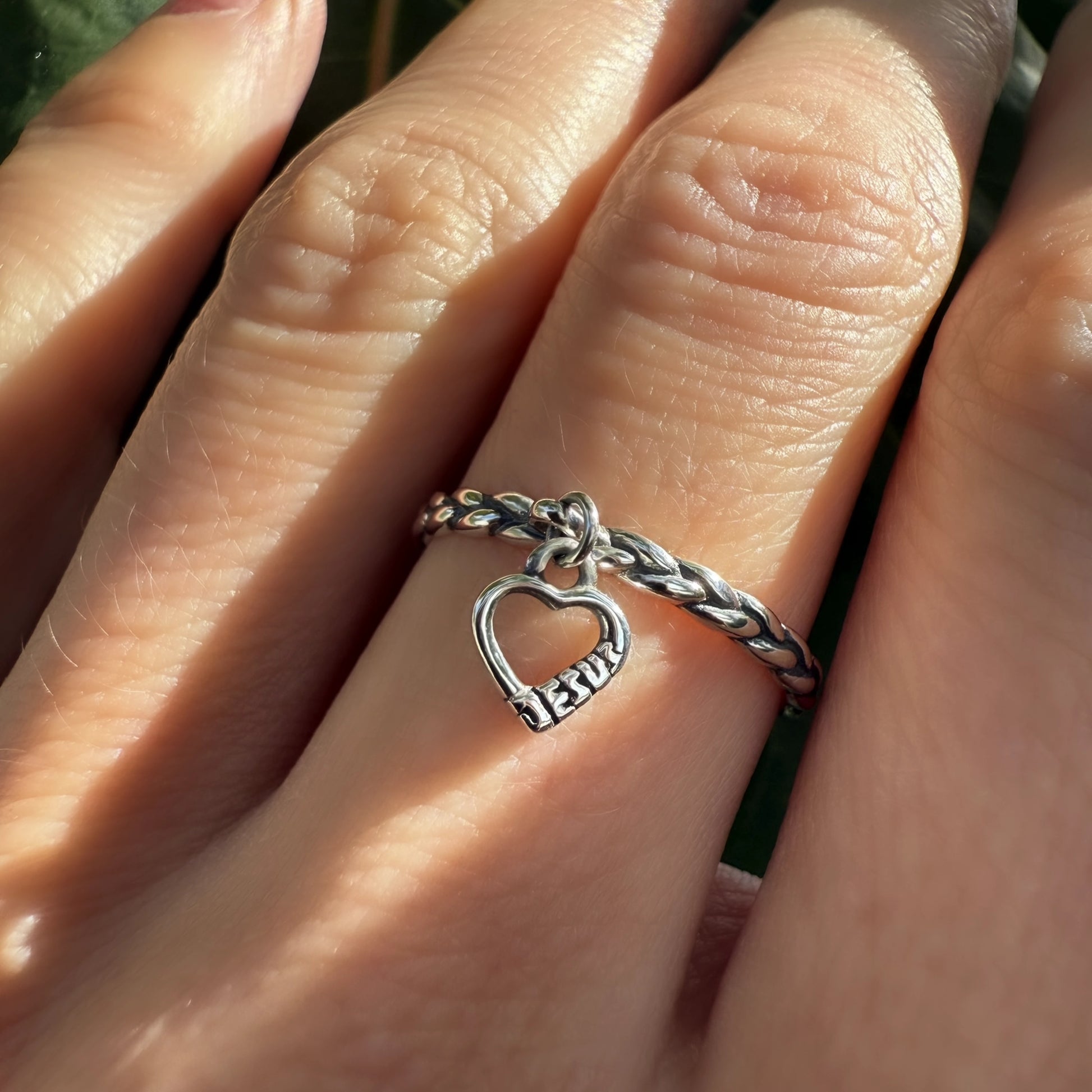 Outdoor shot of a silver heart Jesus ring on the hand, with a blurred leafy background and natural sunlight.