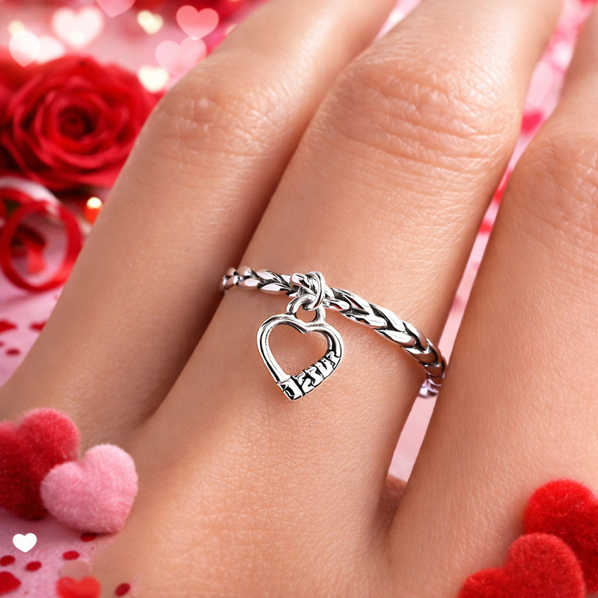 Sterling silver heart charm ring with braided band, worn on a woman’s finger against a romantic Valentine’s Day background with red roses and soft heart bokeh.