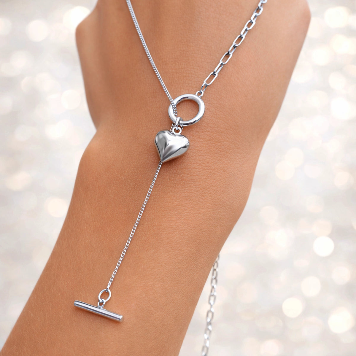 Silver necklace with heart and T-bar pendant on a wrist against a blurred background